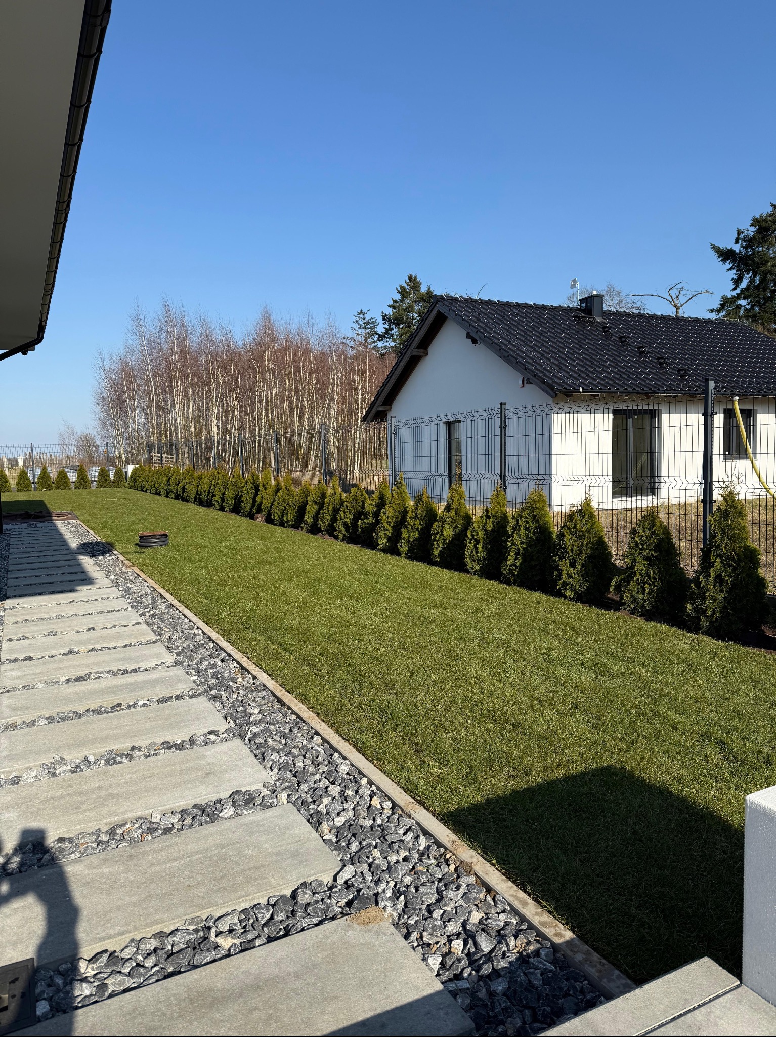 Zadbany trawnik z równo przyciętą trawą, obrzeża z kamienia i ścieżka z płyt betonowych, tuje wzdłuż ogrodzenia. W tle dom z ciemnym dachem i jasną elewacją.