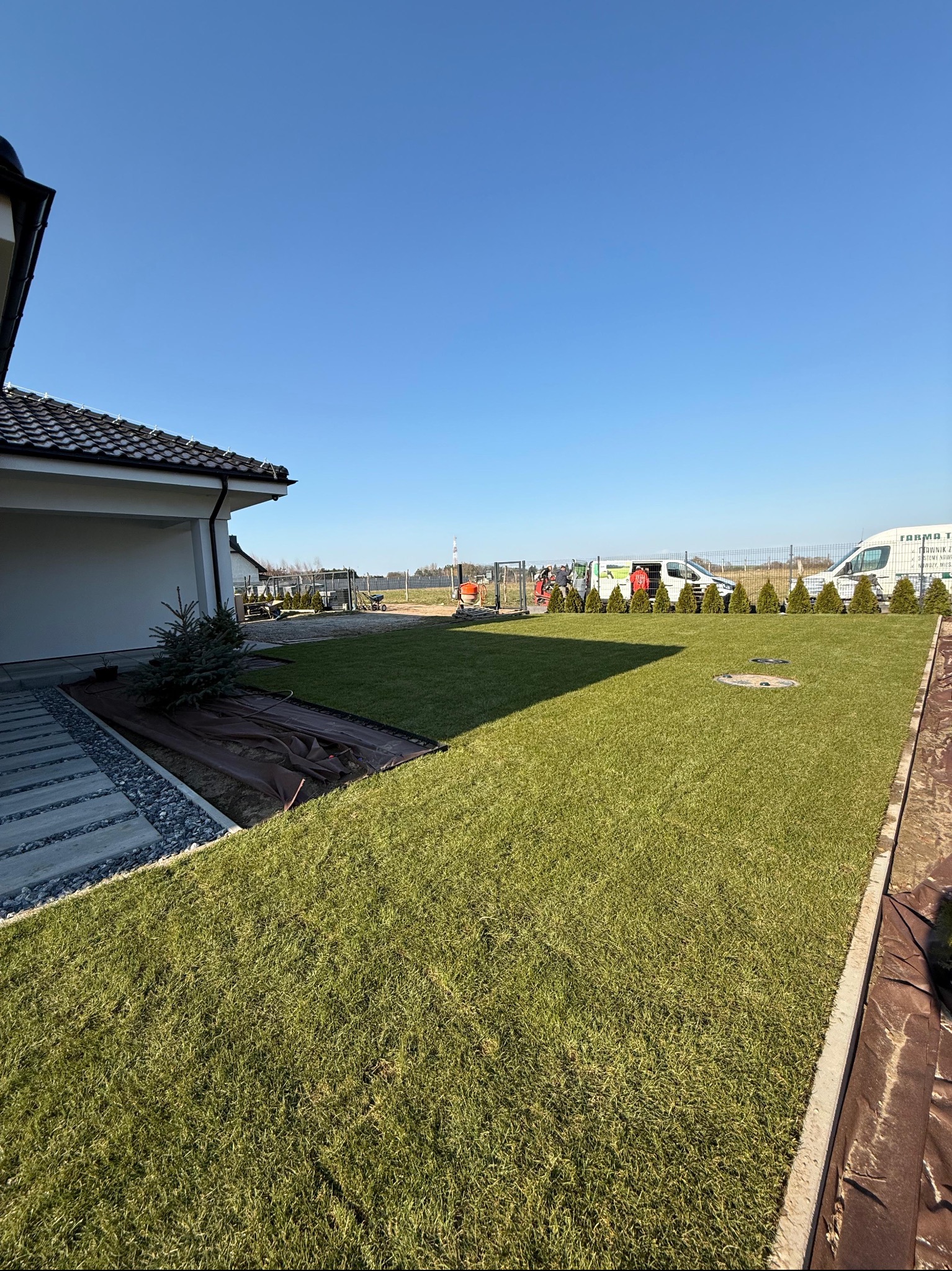 Nowo założony trawnik z rolki przy domu jednorodzinnym, częściowo przykryty brązową tkaniną. W tle widoczne samochody dostawcze i tuje.
