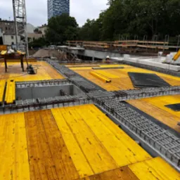 Żółte szalunki i zbrojenia na budowie, widoczni pracownicy w oddali. Konstrukcja w trakcie realizacji, z widocznym fragmentem dźwigu i budynkiem w tle.