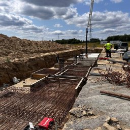T.R.K. BUILDING COMPANY SPÓŁKA Z OGRANICZONĄ ODPOWIEDZIALNOŚCIĄ - Zbrojenie fundamentów pod budynek z użyciem szalunków. Widoczny pracownik i pompa do betonu. W tle wykop i nasyp ziemi. Dzień słoneczny, częściowe zachmurzenie.