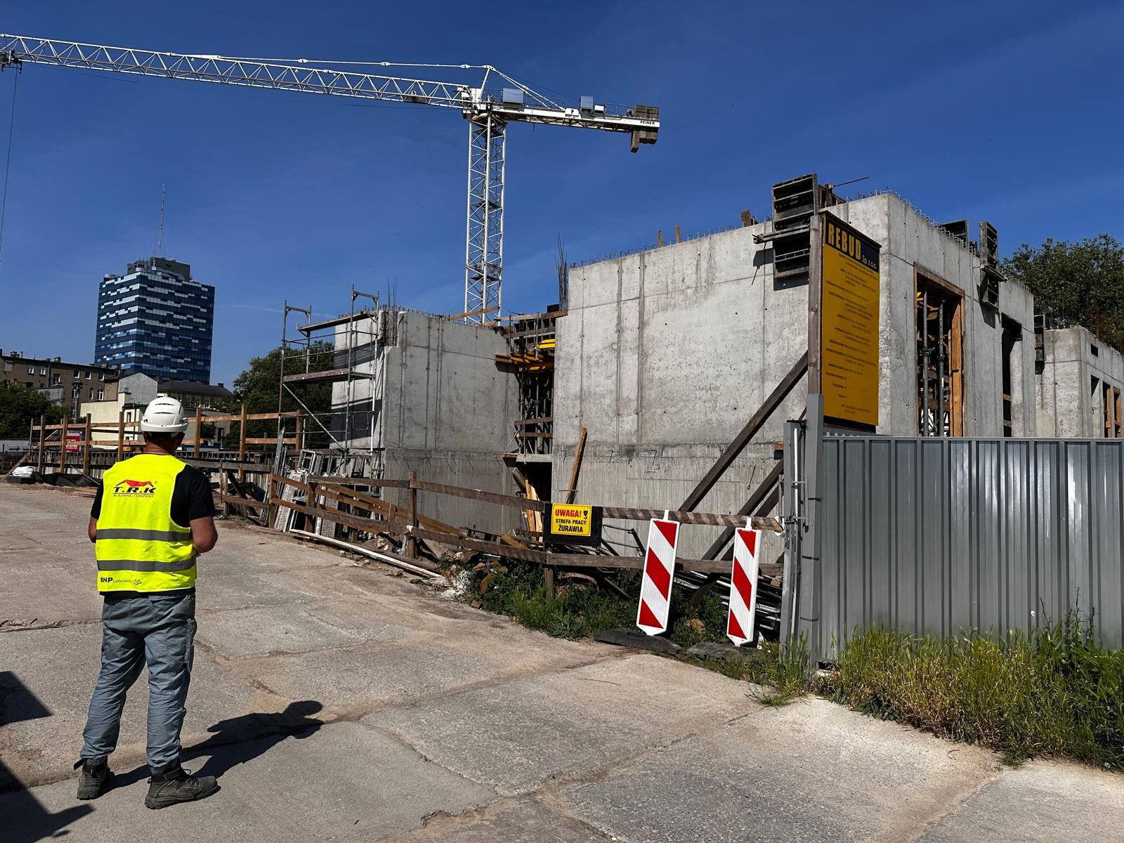 Budowa budynku z betonu: widok na konstrukcję w trakcie wznoszenia, z pracownikiem w kamizelce odblaskowej i żurawiem w tle, w słoneczny dzień.