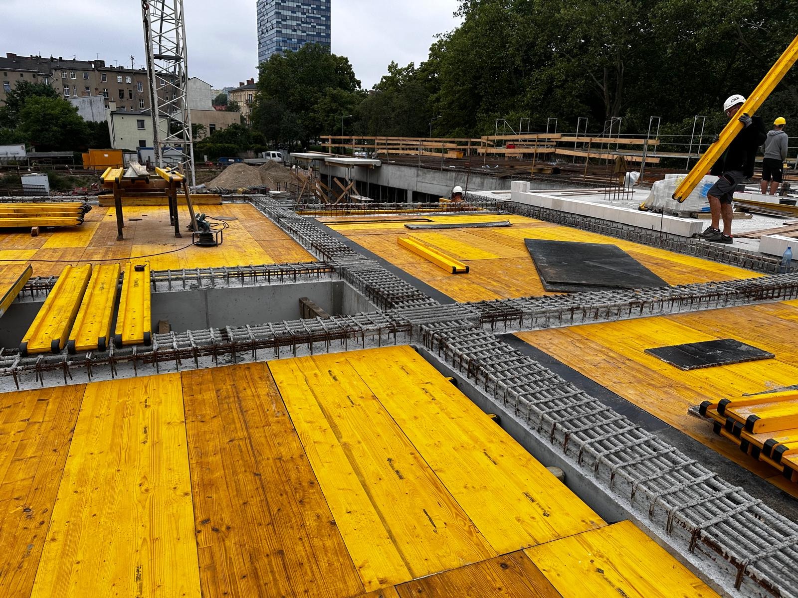 Żółte szalunki i zbrojenia na budowie, widoczni pracownicy w oddali. Konstrukcja w trakcie realizacji, z widocznym fragmentem dźwigu i budynkiem w tle.