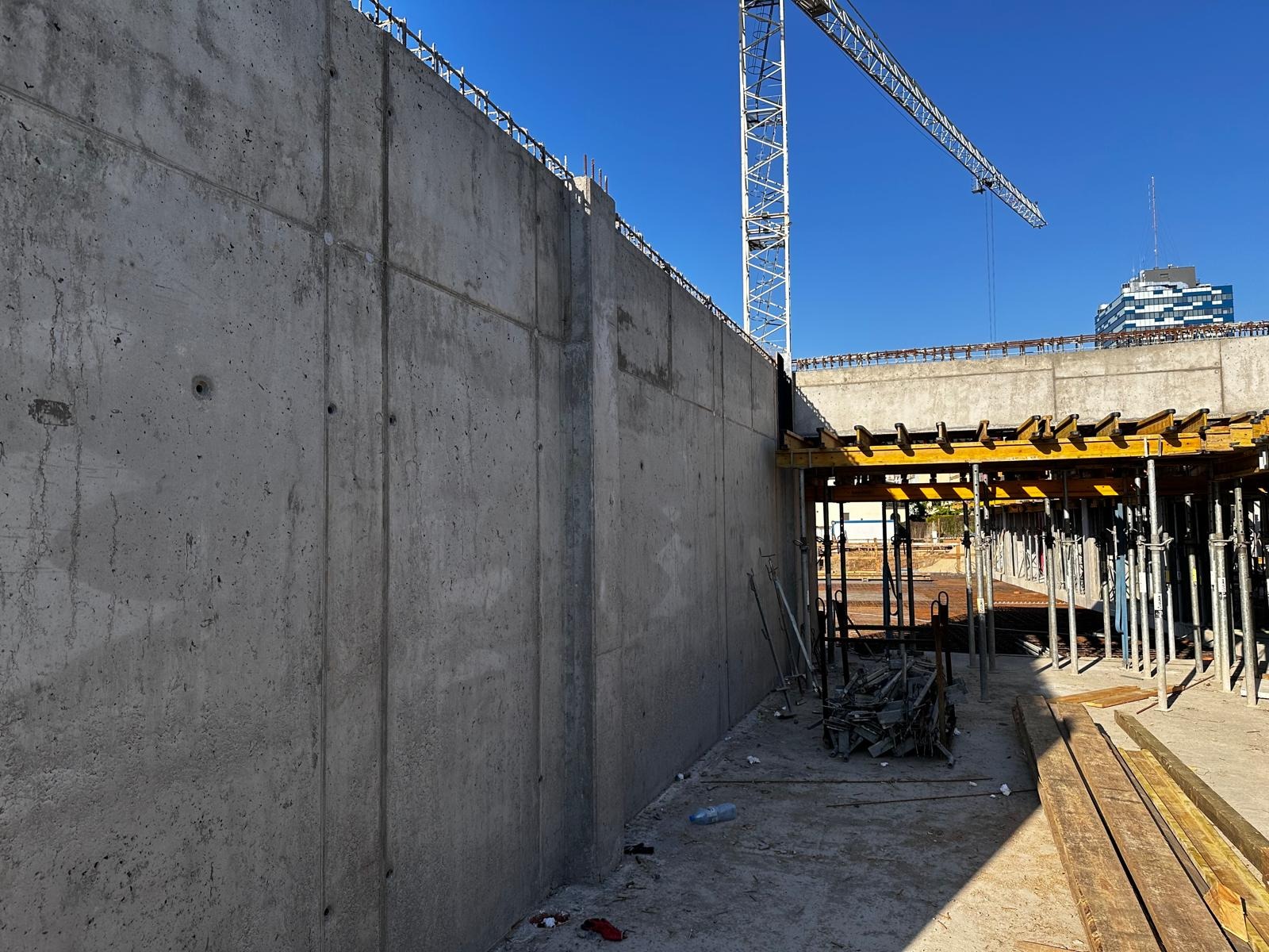 Szara ściana żelbetowa w trakcie budowy, widoczny dźwig budowlany i szalunki. Elementy zbrojenia na górze ściany. Budynek w tle. Słoneczny dzień.