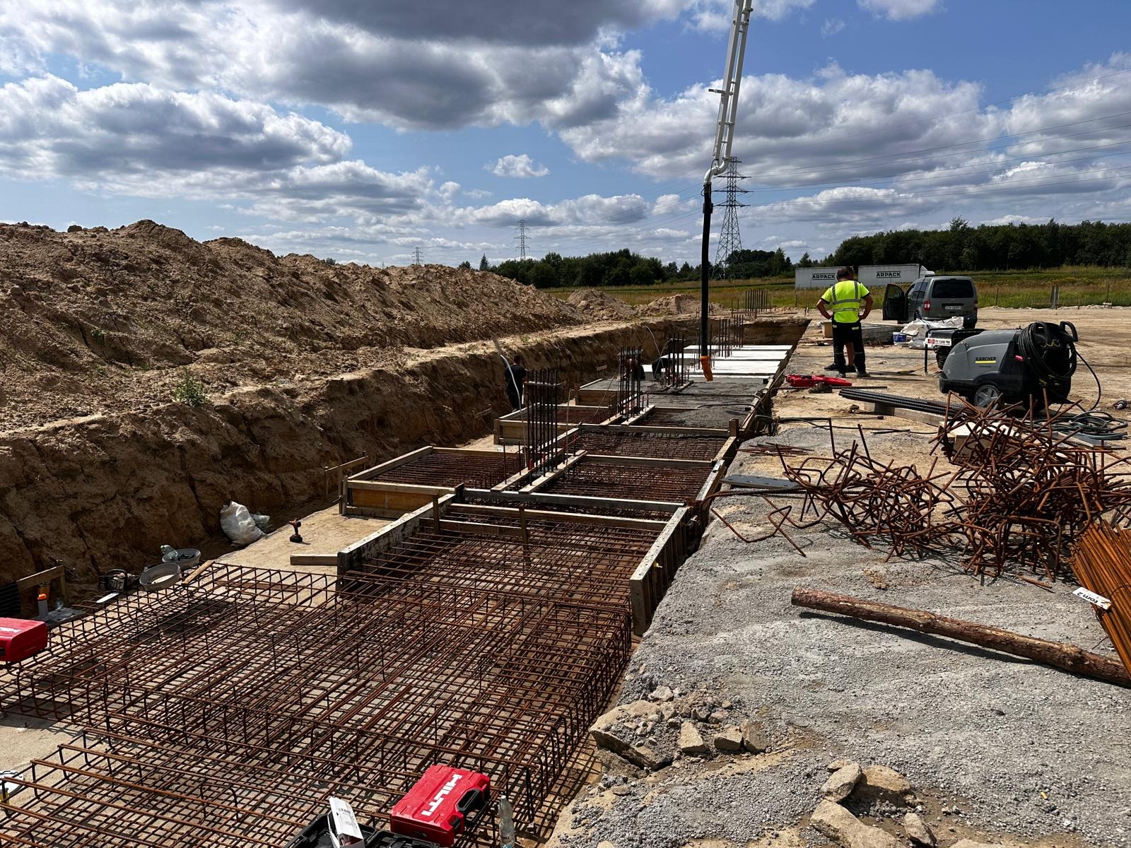 Zbrojenie fundamentów pod budynek z użyciem szalunków. Widoczny pracownik i pompa do betonu. W tle wykop i nasyp ziemi. Dzień słoneczny, częściowe zachmurzenie.