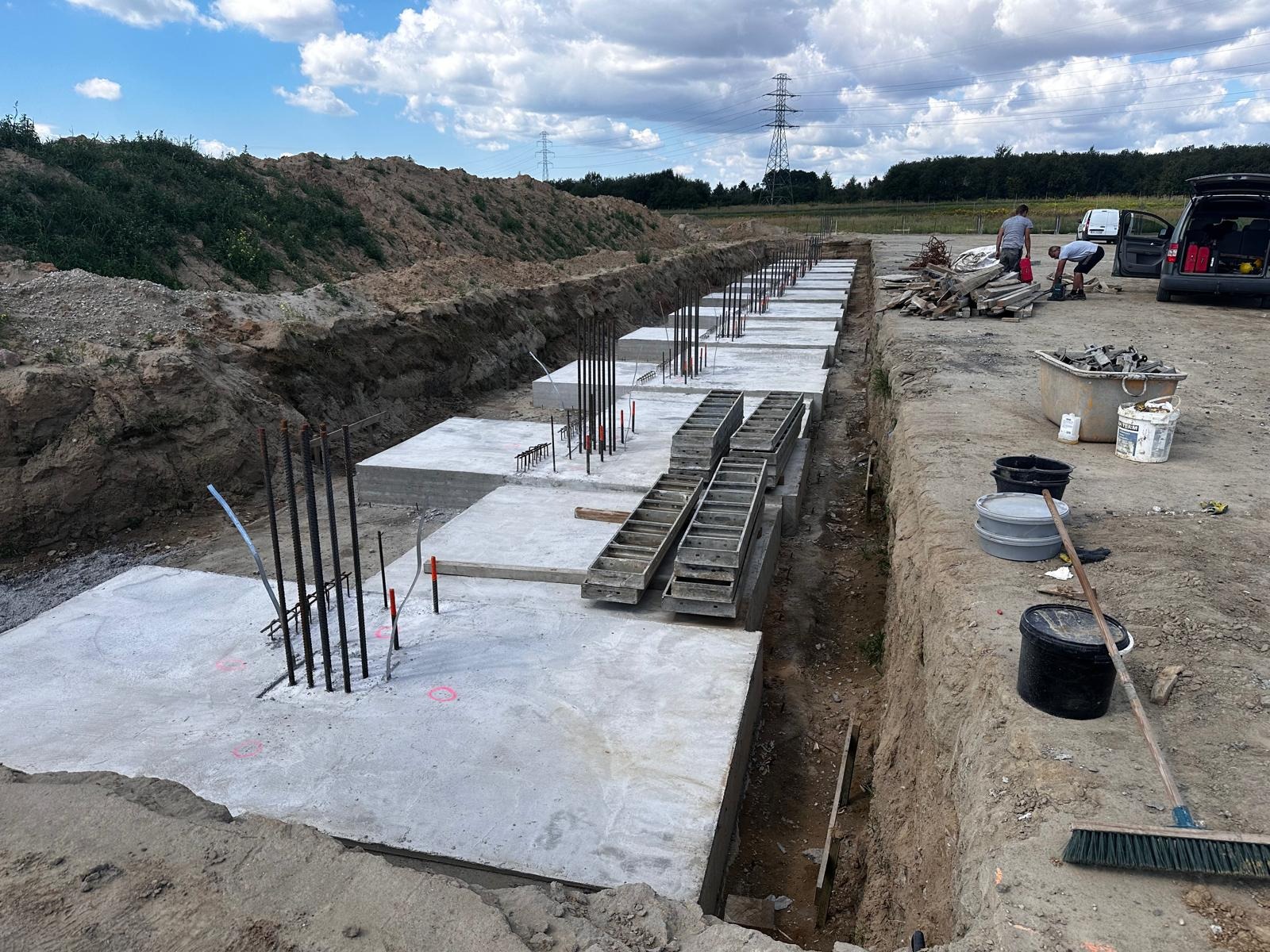 Wykop pod fundamenty z częściowo zalanymi betonem stopami fundamentowymi i wystającymi prętami zbrojeniowymi. Na drugim planie widoczni pracownicy i samochód.