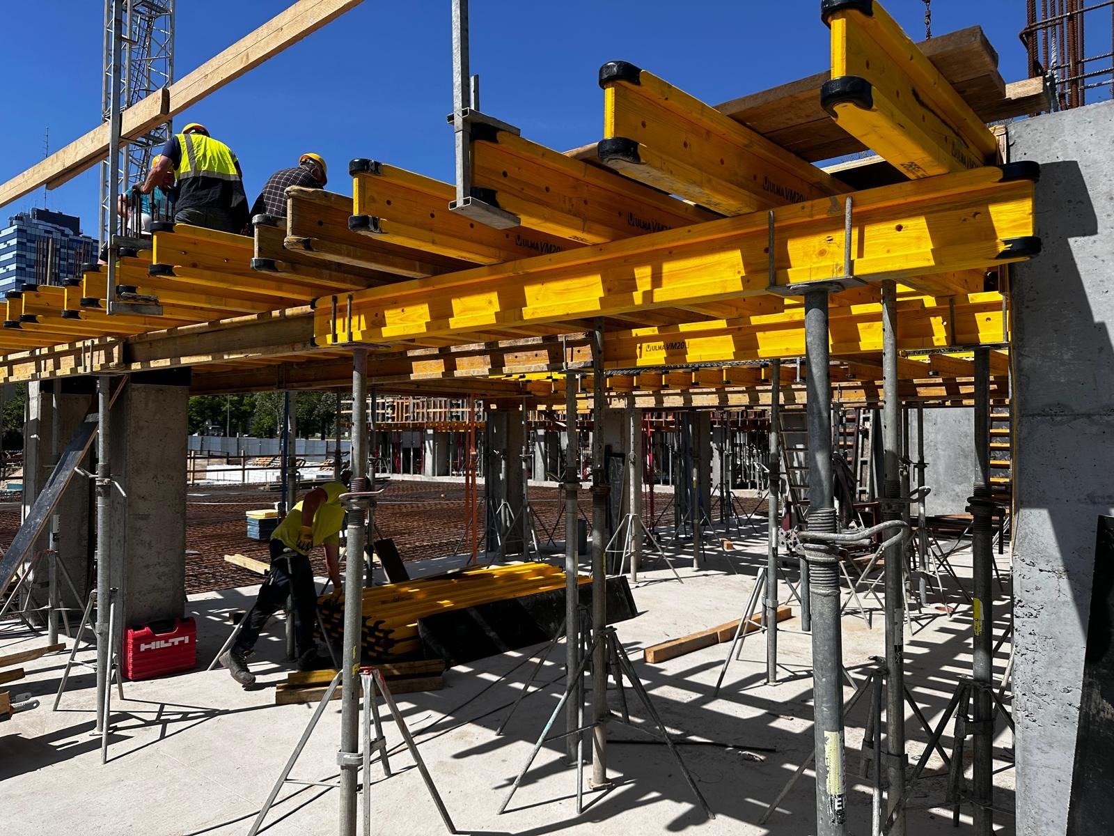 Budowa budynku z żelbetu: pracownicy montują żółte szalunki stropowe na stalowych podporach. Widoczne zbrojenia i narzędzia Hilti. Słoneczny dzień.