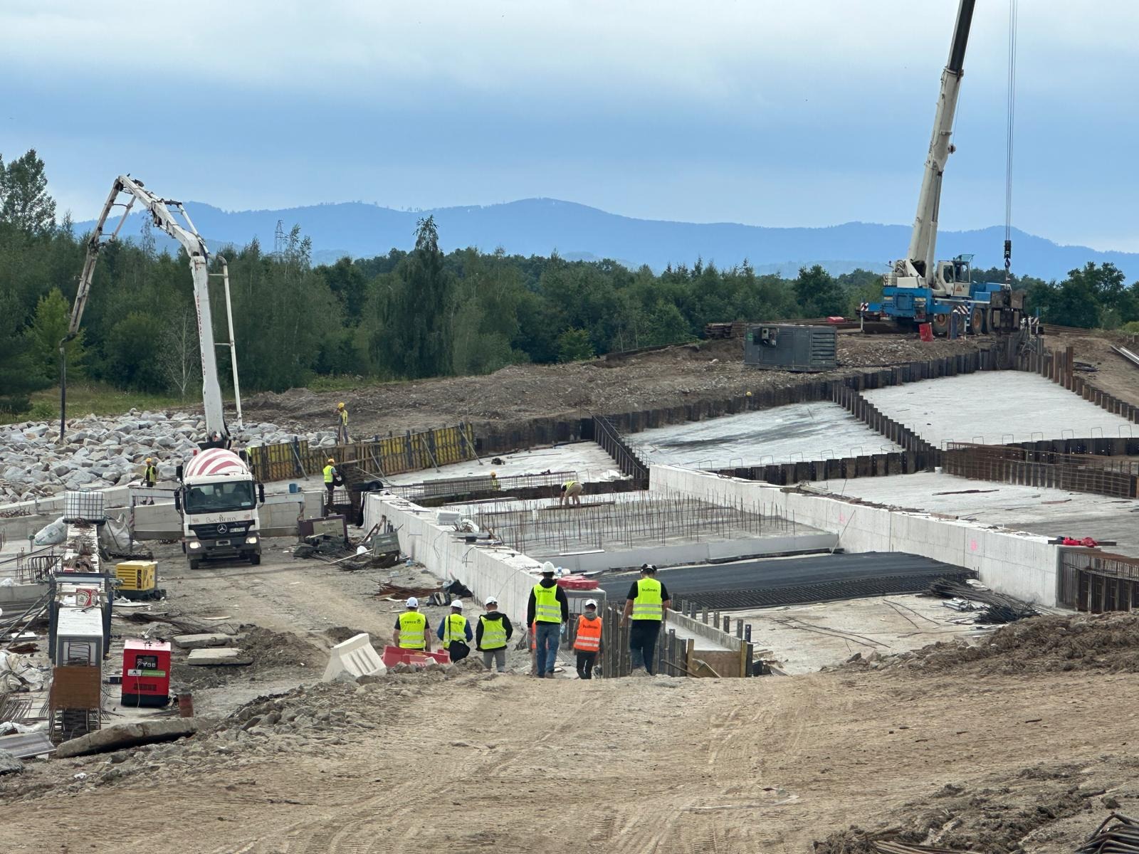 Budowa fundamentów: ekipa w kamizelkach nadzoruje zbrojenie i betonowanie. W tle betoniarka i żuraw, a w oddali zalesione wzgórza.