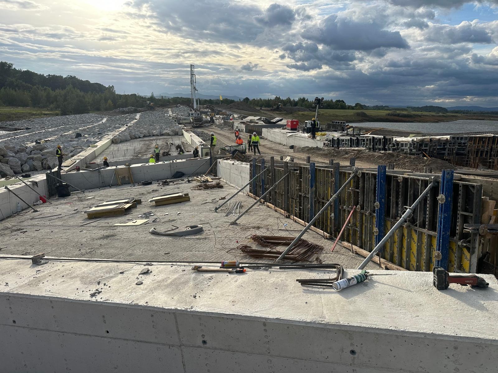Budowa żelbetowej konstrukcji nad wodą. Widoczne szalunki, zbrojenia, betonowe elementy i pracownicy w odblaskowych kamizelkach. W tle krajobraz z jeziorem i lasem.