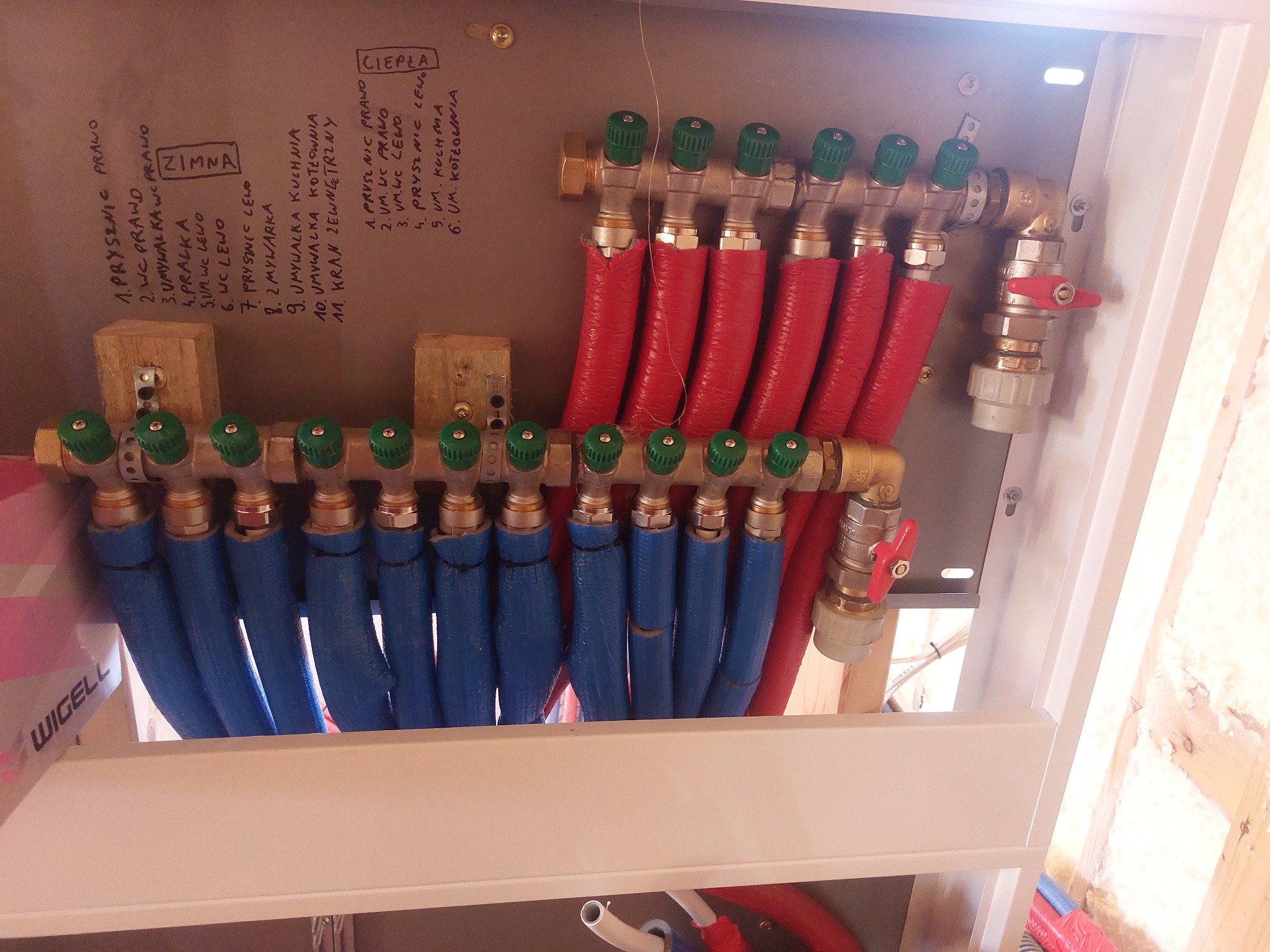 Rozdzielacz ogrzewania podłogowego w szafce, z rurami w izolacji czerwonej i niebieskiej, opisany odręcznie listą punktów poboru wody.