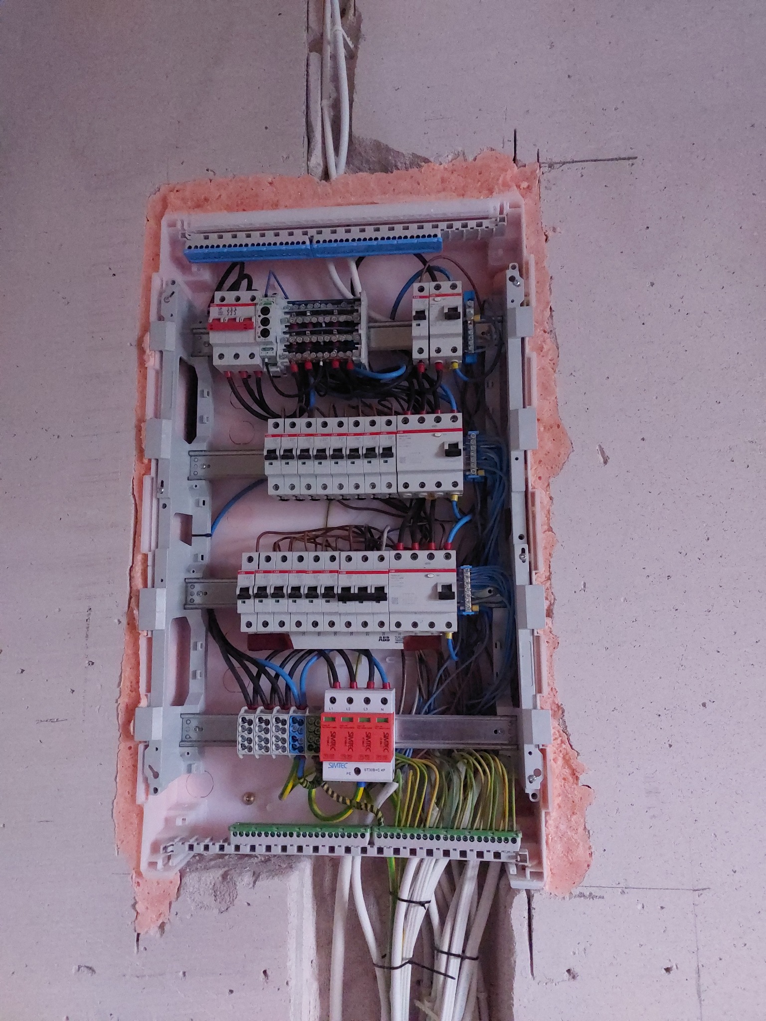 Rozdzielnica elektryczna w trakcie montażu, widoczne zabezpieczenia, przewody i szyny DIN. Ściana w surowym stanie, obudowa rozdzielnicy zabezpieczona pianką montażową.