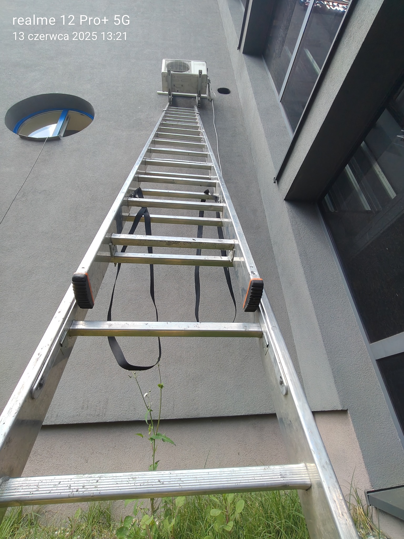 Długa drabina oparta o szarą ścianę budynku, prowadząca do zamontowanego klimatyzatora. Nietypowe okrągłe okno w tle. Widok z dołu, trawa u podstawy drabiny.