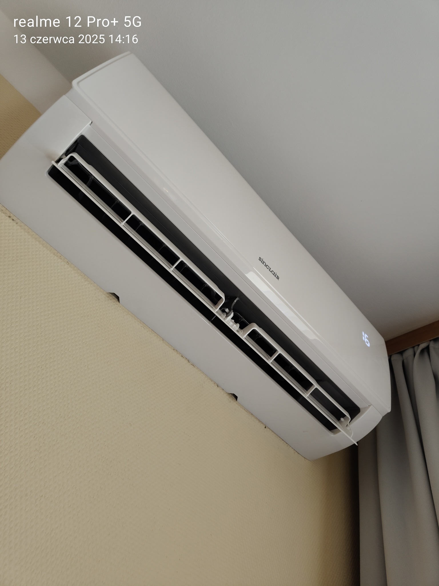 Biała klimatyzacja Sinclair zamontowana na ścianie z beżową tapetą, widoczny wyświetlacz temperatury i fragment zasłony. Ujęcie z dołu.