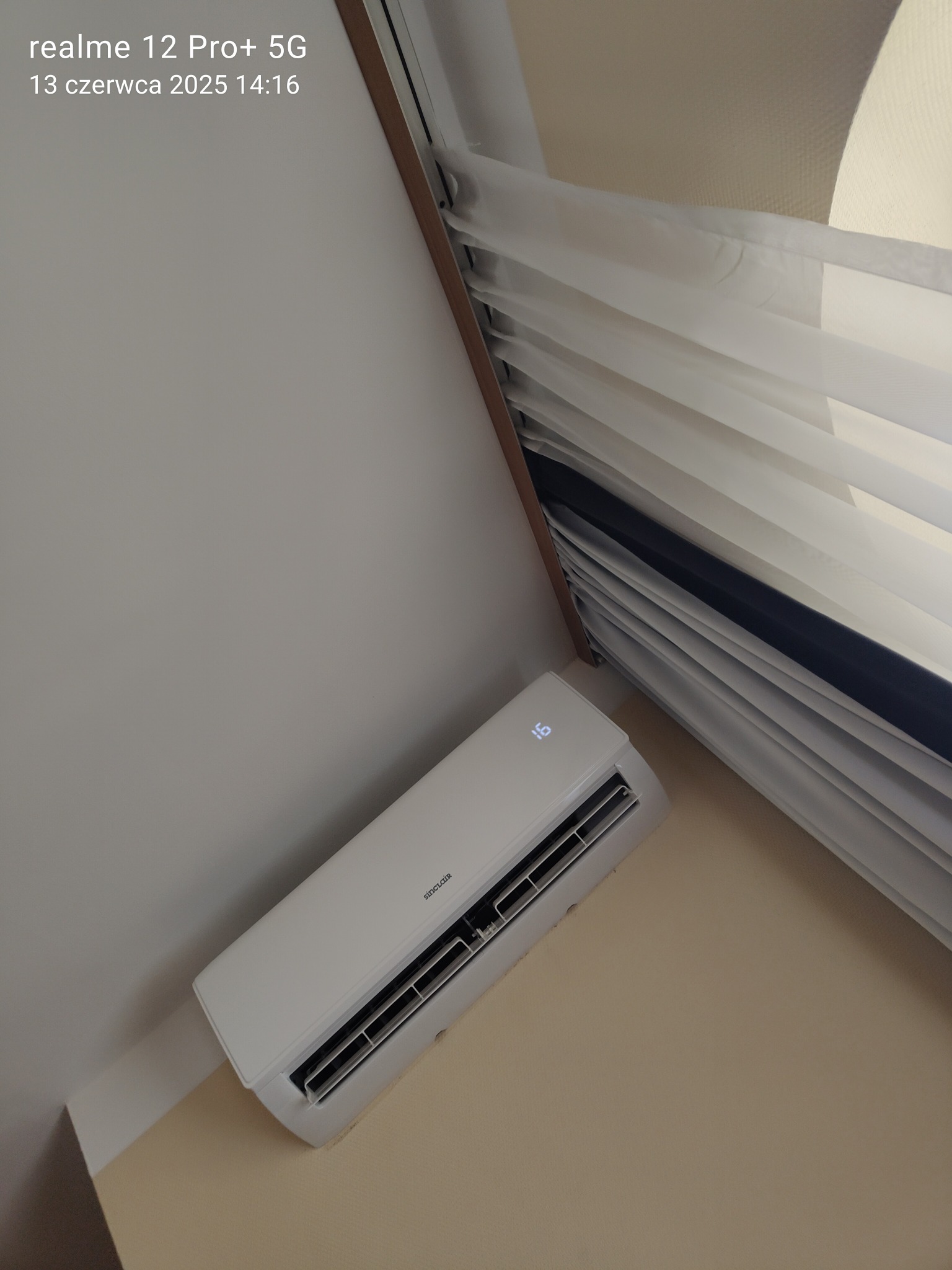 Biała klimatyzacja Sinclair zamontowana na ścianie w pomieszczeniu z beżowymi ścianami, widok z dołu, obok okna z żaluzjami.
