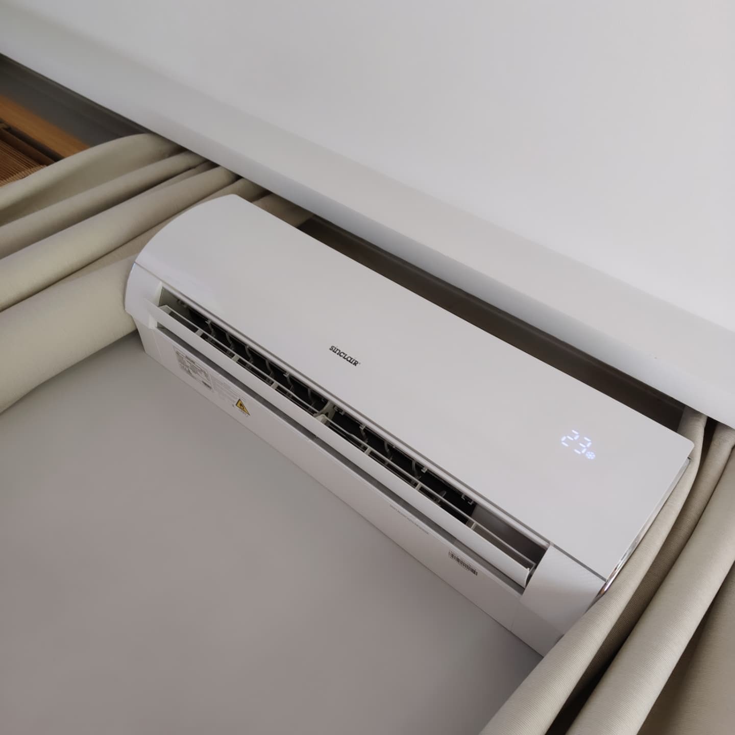 Biała klimatyzacja Sinclair na ścianie, widoczny wyświetlacz z temperaturą 23 stopnie, zasłonięta częściowo zasłoną. Montaż klimatyzacji w domu.