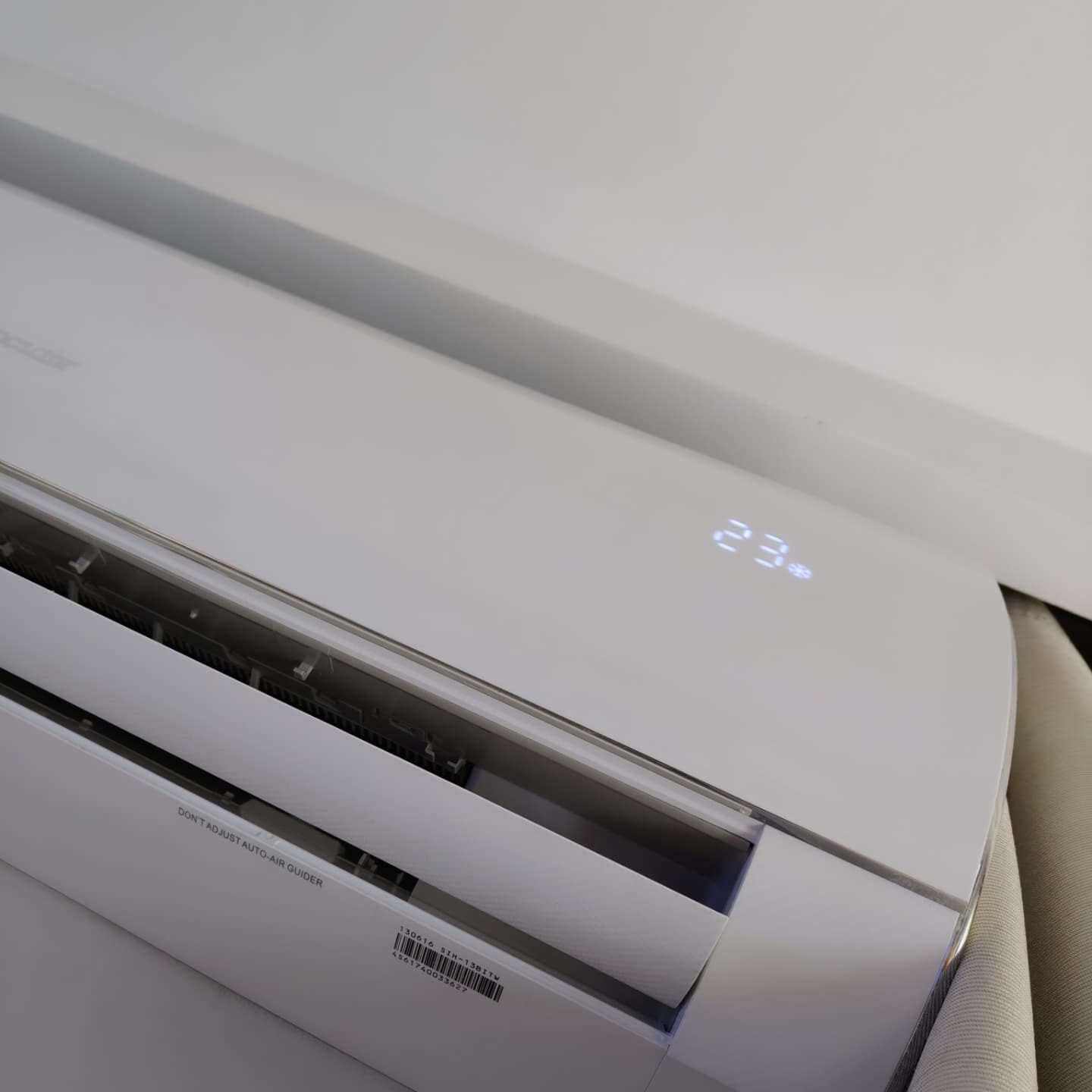 Biała klimatyzacja ścienna z wyświetlaczem temperatury 23°C, widoczna z bliska pod kątem, z fragmentem ściany w tle.