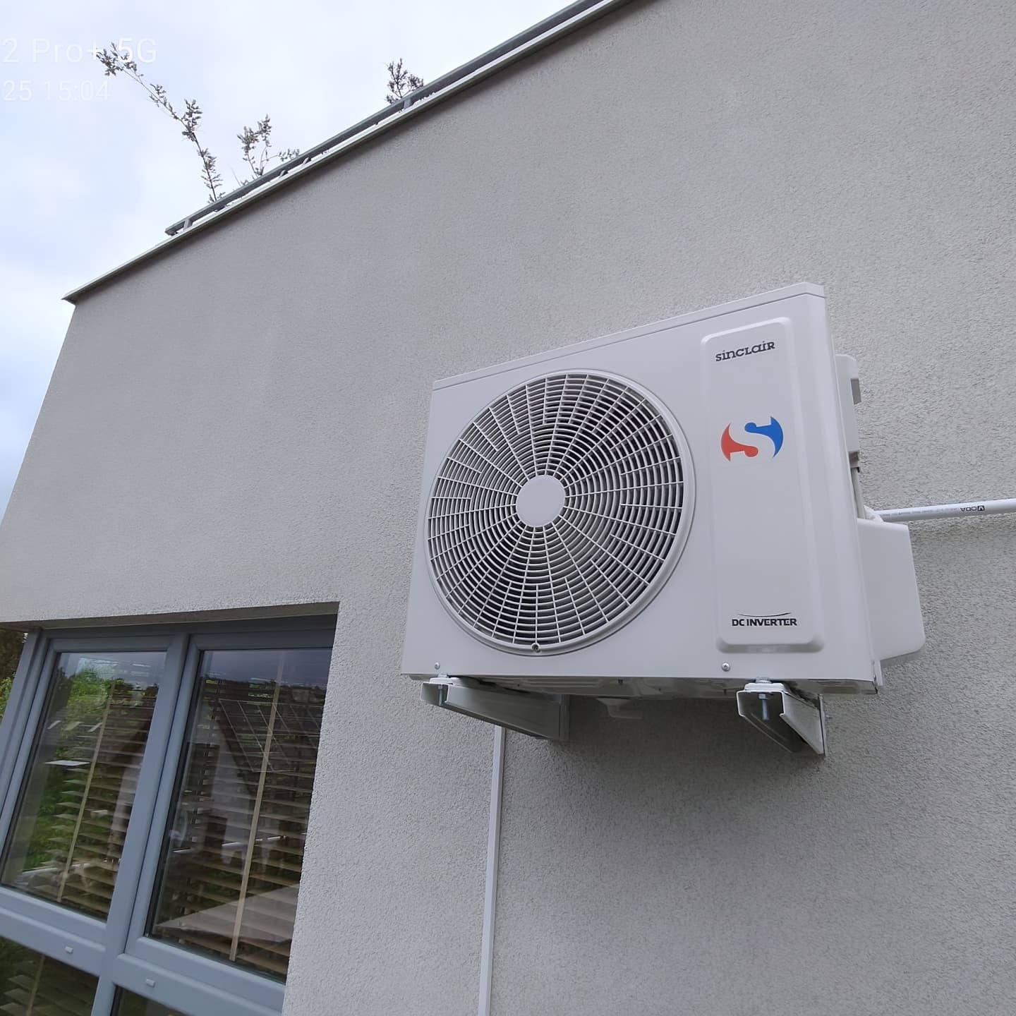 Biała jednostka zewnętrzna klimatyzacji Sinclair, zamontowana na jasnej elewacji budynku z widocznym oknem. Minimalistyczny design, nowoczesny wygląd.