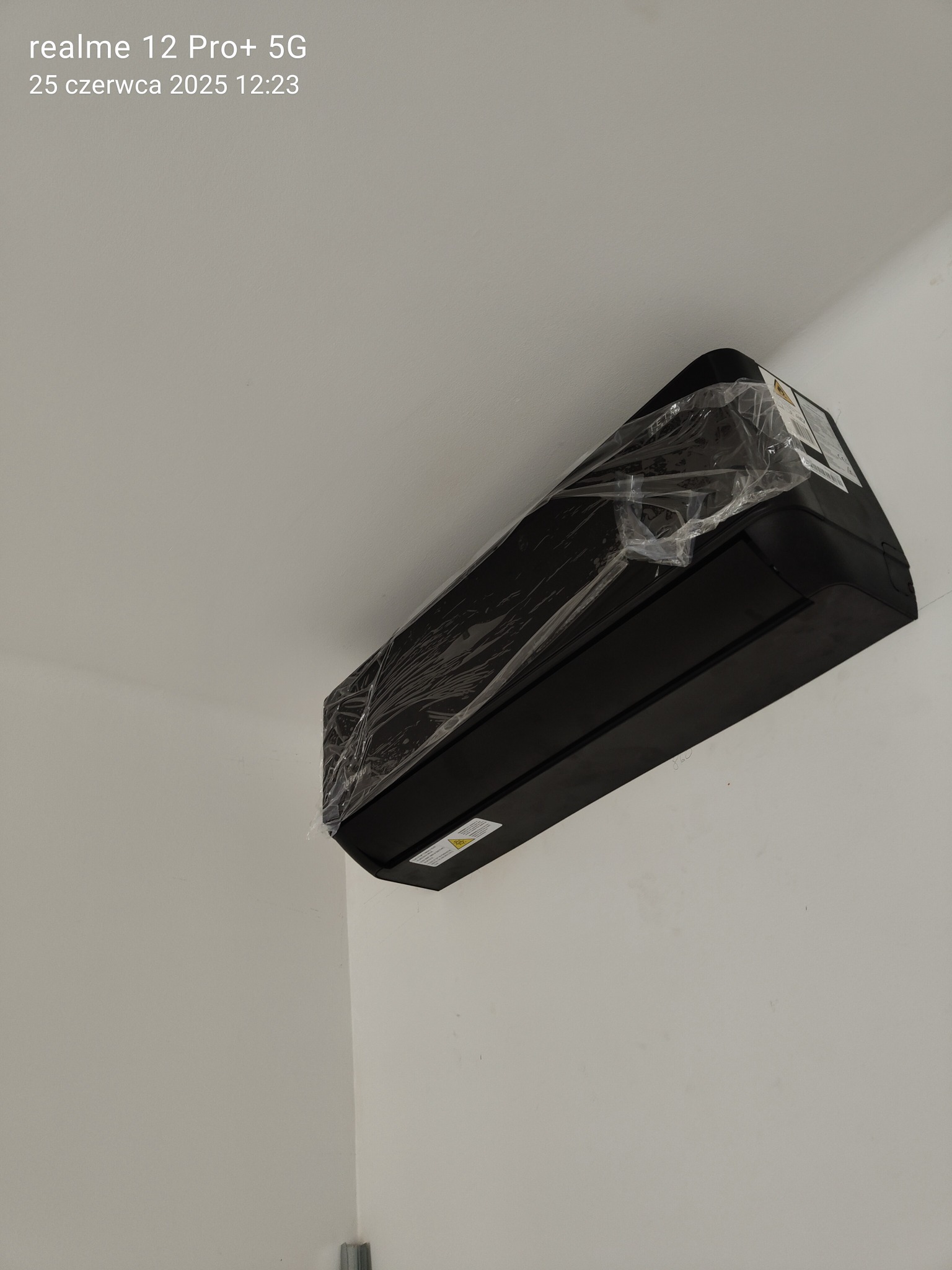 Czarna jednostka wewnętrzna klimatyzacji, jeszcze w folii ochronnej, zamontowana na białej ścianie. Widoczne detale montażu.