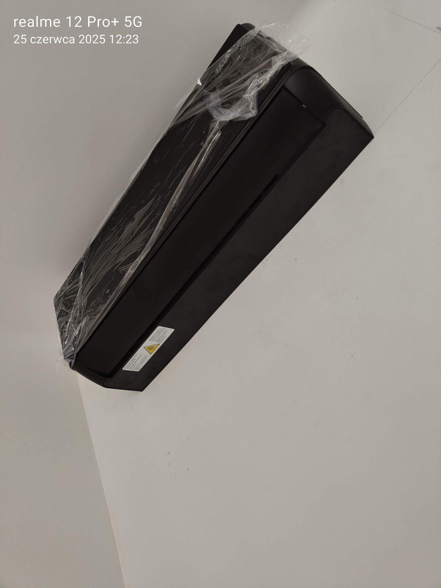 Nowy, czarny klimatyzator ścienny, jeszcze w folii ochronnej, zamontowany na jasnej ścianie. Widoczna naklejka ostrzegawcza na urządzeniu. Lekki kąt ujęcia.