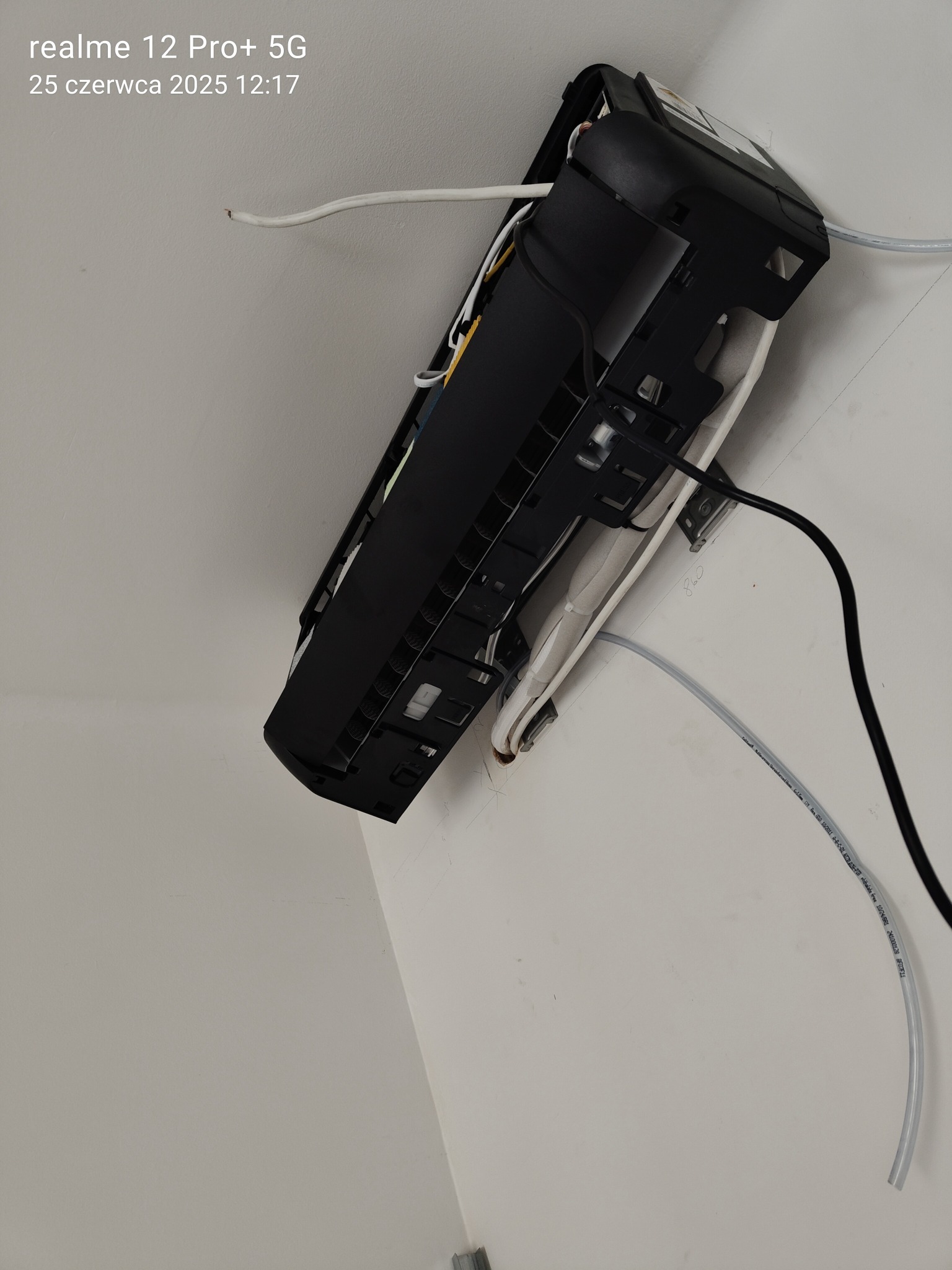 Czarny klimatyzator ścienny w trakcie montażu, widoczne przewody i rurki instalacyjne na białej ścianie. Ujęcie z dołu, z widocznymi elementami mocującymi.