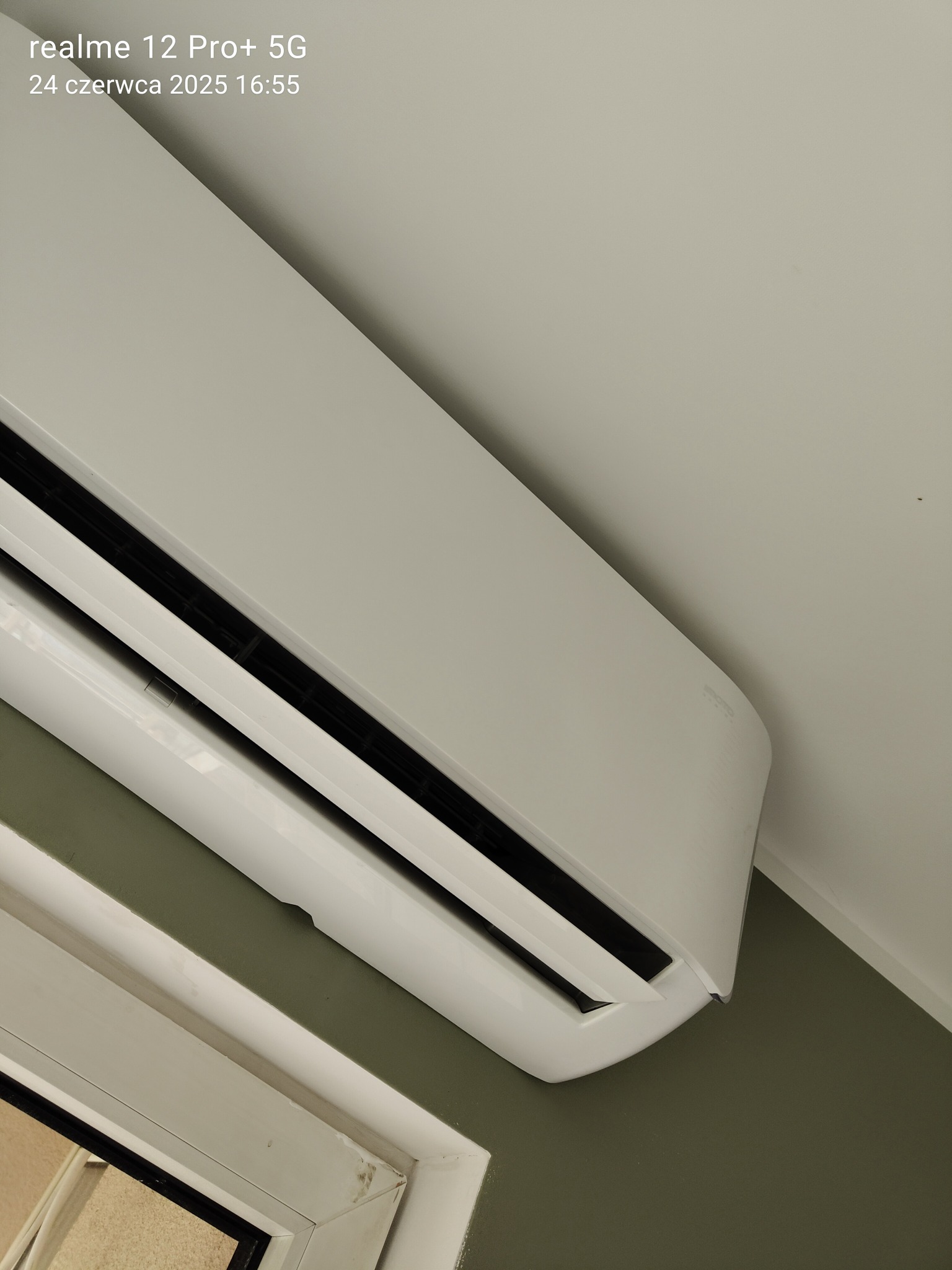 Biała klimatyzacja ścienna zamontowana w rogu pokoju z zieloną ścianą, widoczny fragment okna. Ujęcie z dołu, z widocznym modelem klimatyzatora.