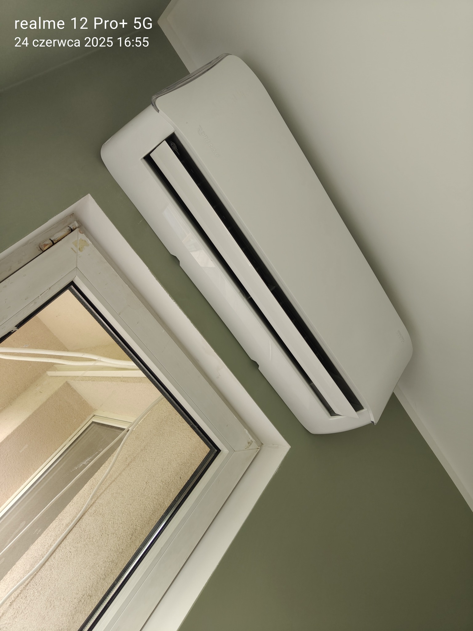 Biały klimatyzator Rotenso, zamontowany na ścianie obok okna z widocznymi kablami na zewnątrz. Ujęcie z dołu, lekko zniekształcona perspektywa.