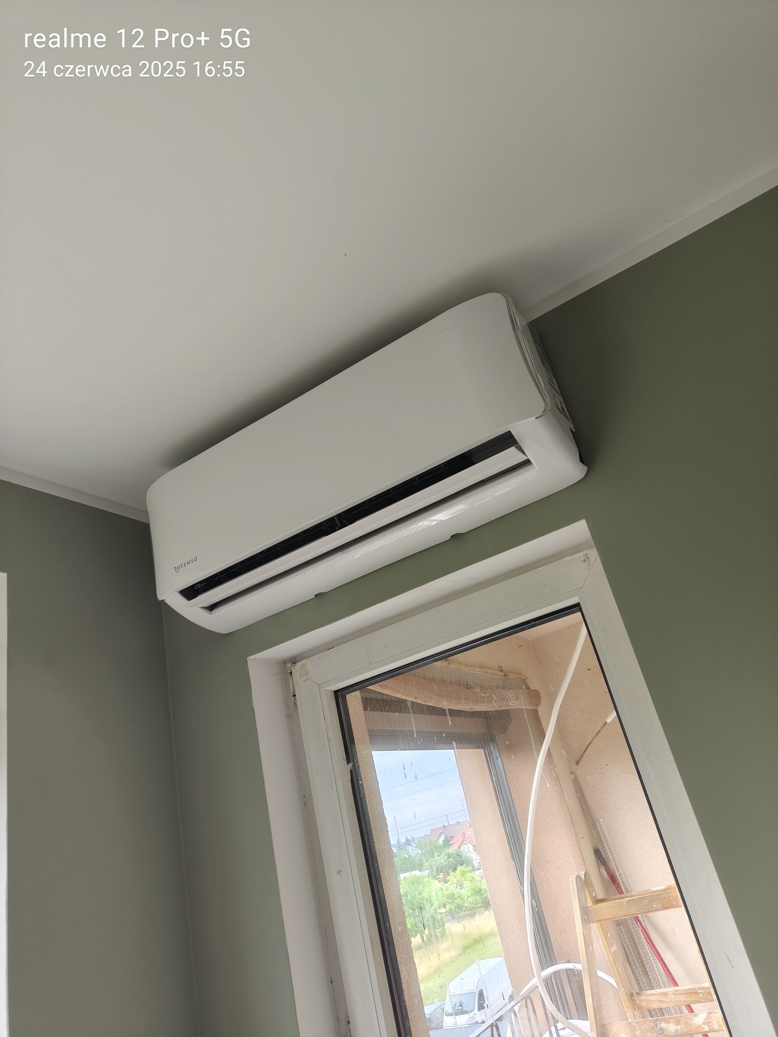 Biała klimatyzacja Rotenso zamontowana na ścianie w pokoju, widok z dołu, obok okno z widokiem na balkon i drabinę. Ściana pomalowana na oliwkowy kolor.