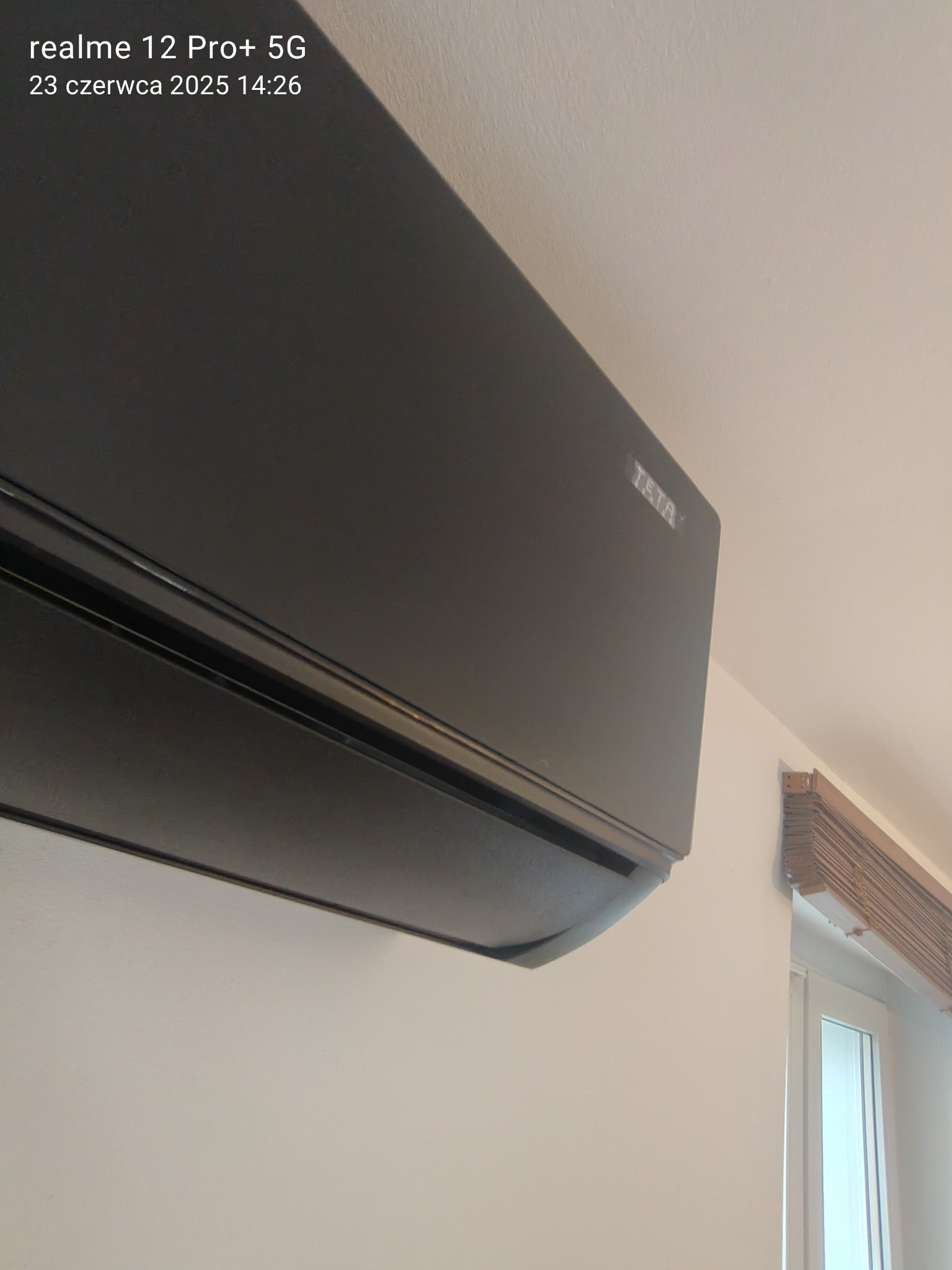 Czarny klimatyzator TATA zamontowany na białej ścianie, widok z dołu, w tle fragment okna z drewnianymi żaluzjami. Nowoczesny design.