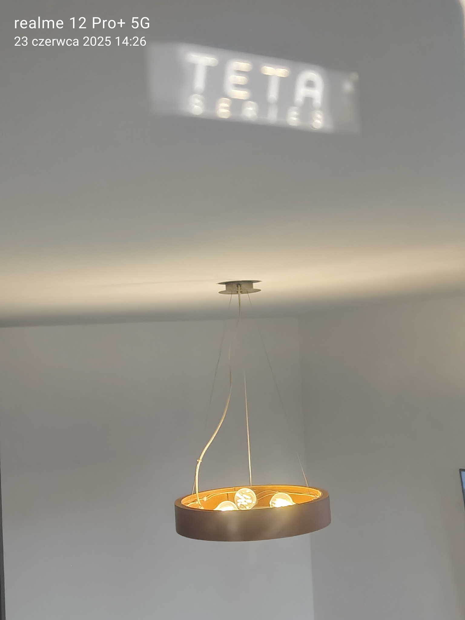 Nowoczesny okrągły żyrandol z ciepłym światłem, wiszący na tle białego sufitu z widocznym odbiciem napisu 'TETA SERIES'.