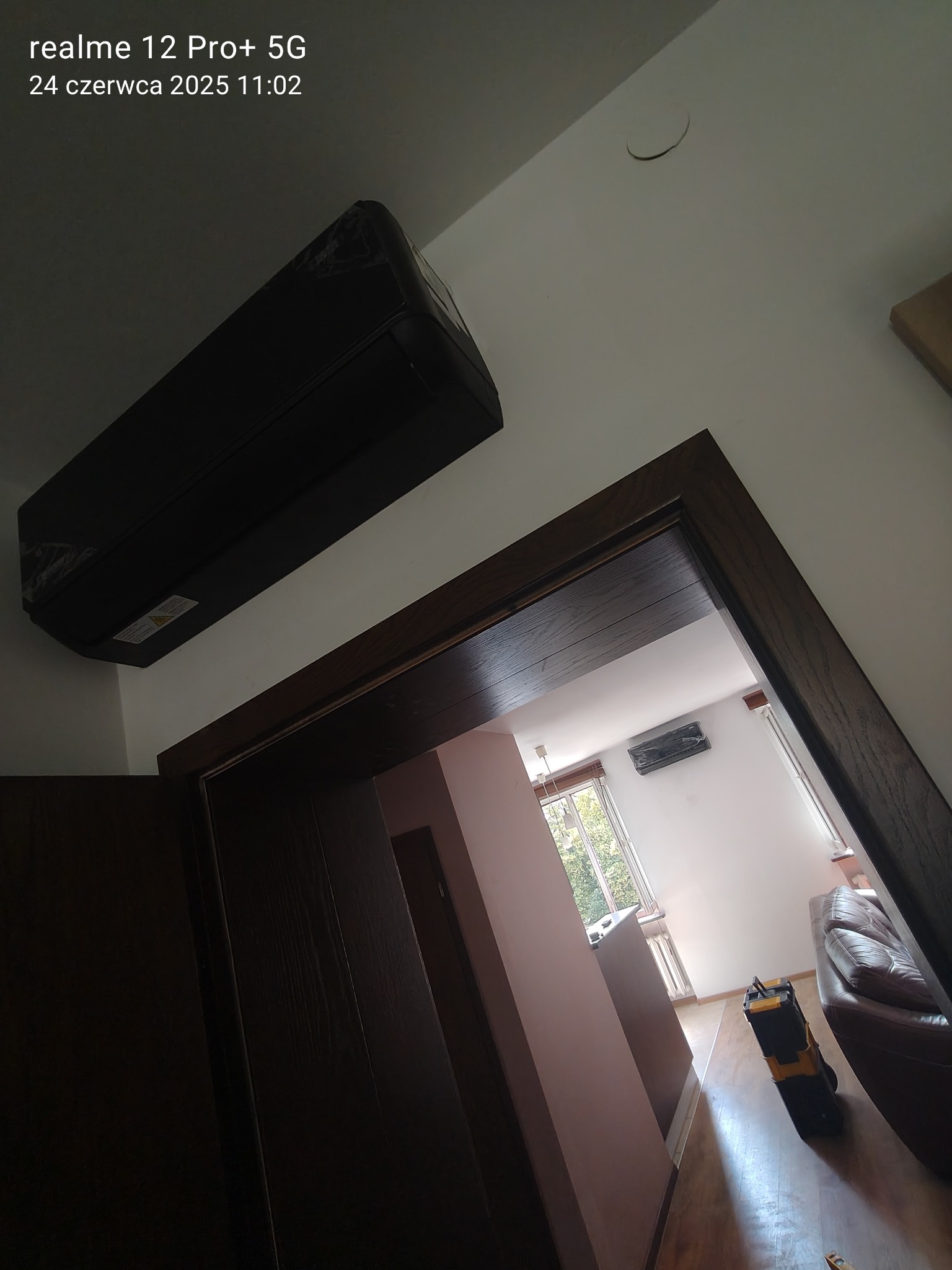 Czarny klimatyzator ścienny w folii ochronnej, widoczny zza drewnianej futryny drzwi, z narzędziami w pokoju w tle, sfotografowany z niskiego kąta.