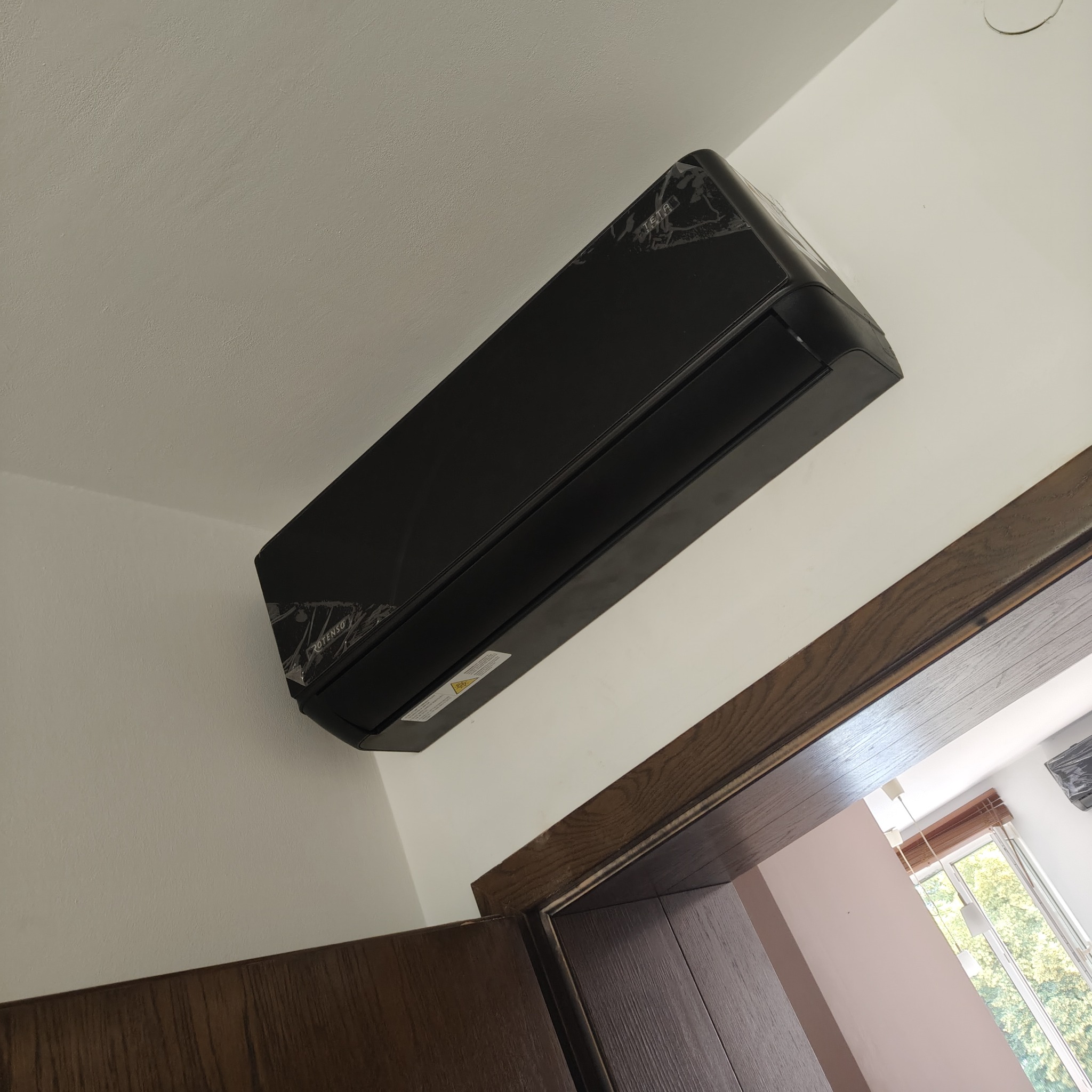 Czarny klimatyzator na ścianie, jeszcze w folii ochronnej, widoczny fragment futryny drzwi i okno w tle. Widok z dołu.