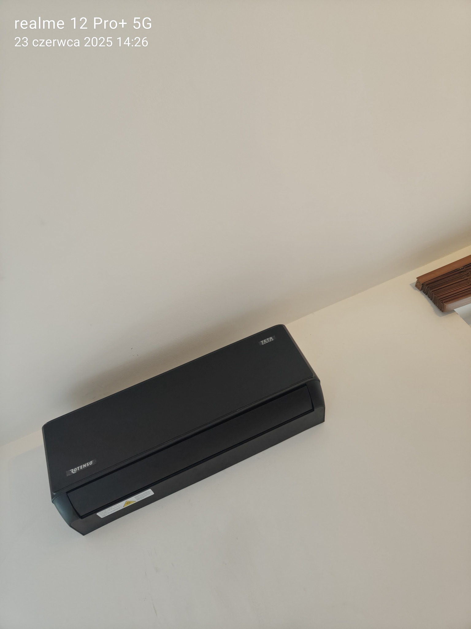 Czarny klimatyzator Rotenso zamontowany na jasnej ścianie. Widoczny fragment żaluzji w prawym górnym rogu. Ujęcie z dołu, lekko z prawej strony.