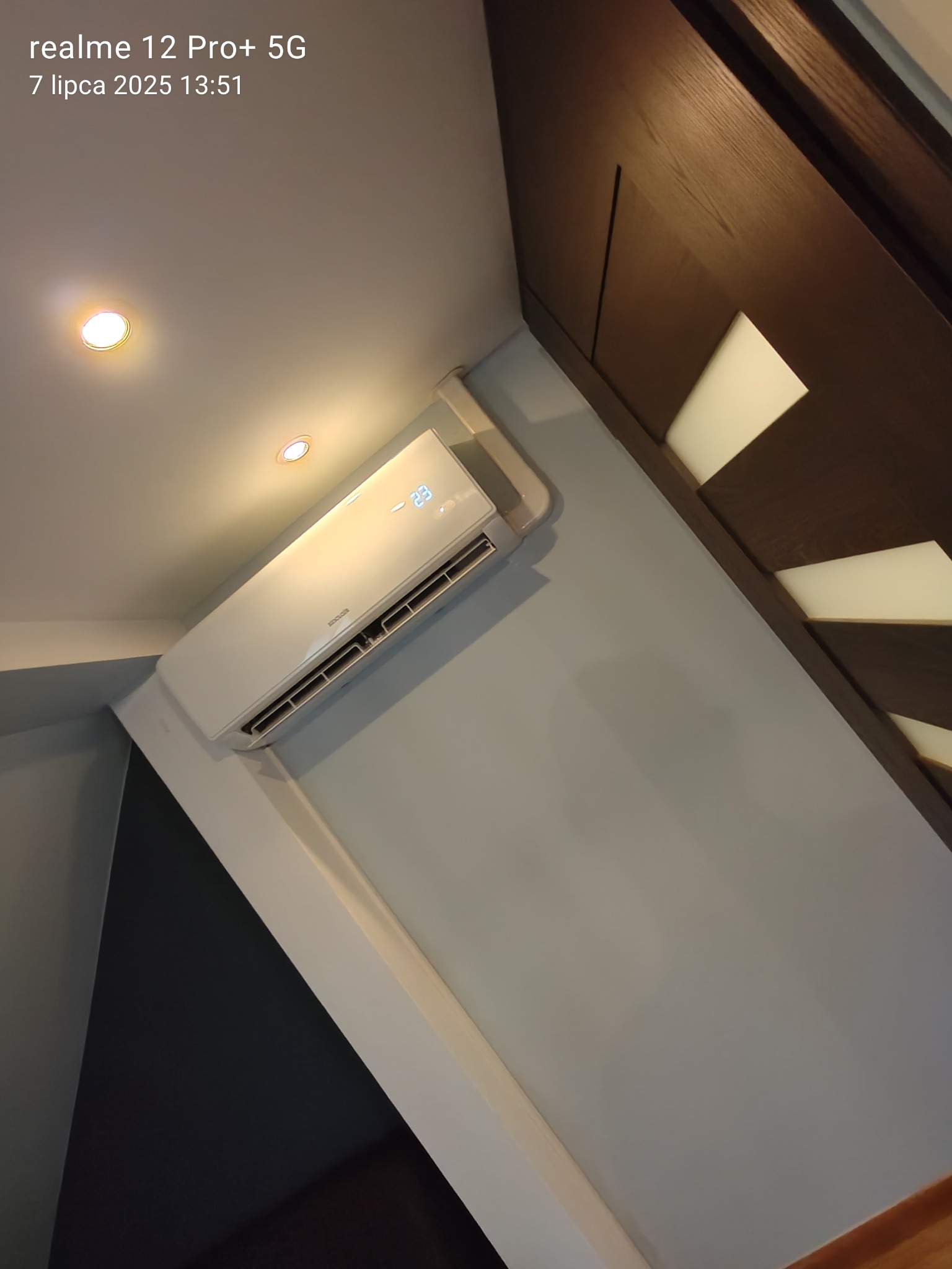 Biała klimatyzacja ścienna na szarej ścianie, widoczny fragment drewnianej zabudowy. Ustawiona temperatura 23 stopnie Celsjusza, oświetlenie sufitowe.