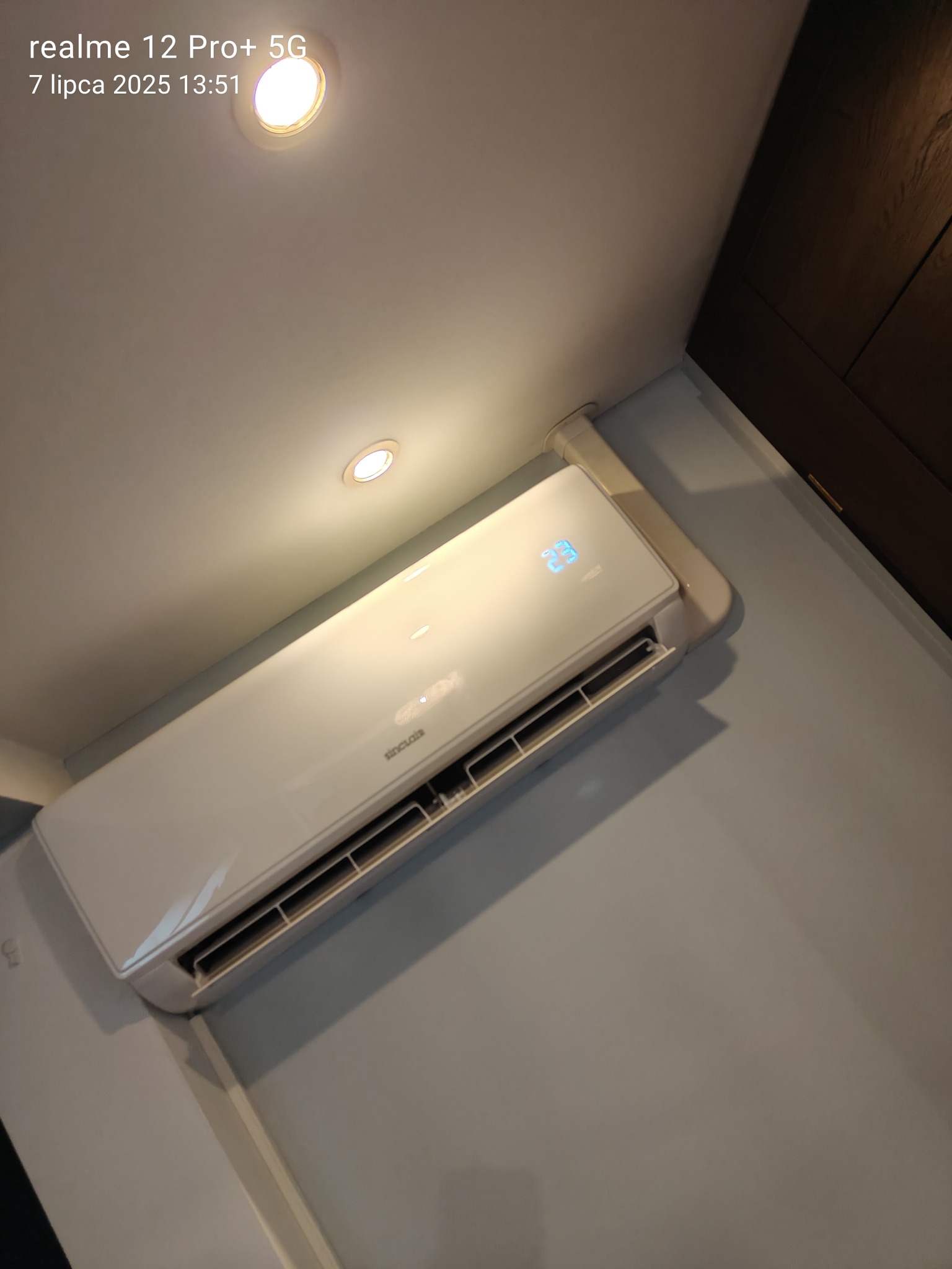 Biały klimatyzator Sinclaire z wyświetlaczem 23°C, zamontowany na jasnej ścianie w rogu pokoju, oświetlony punktowym światłem z sufitu.