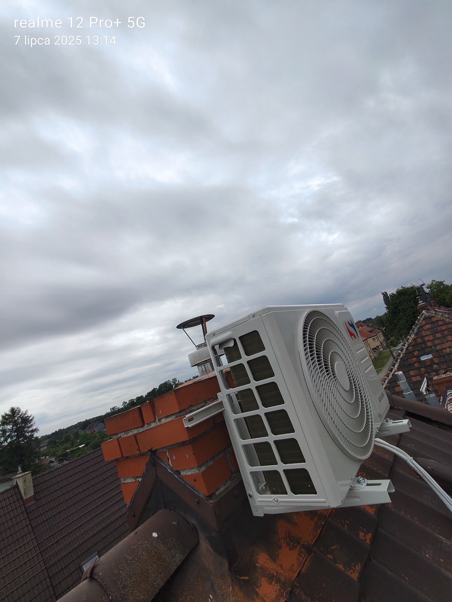 Jednostka zewnętrzna klimatyzacji zamontowana na dachu, z widocznym uchwytem i przewodami. Tło stanowi pochmurne niebo i zabudowa mieszkalna.