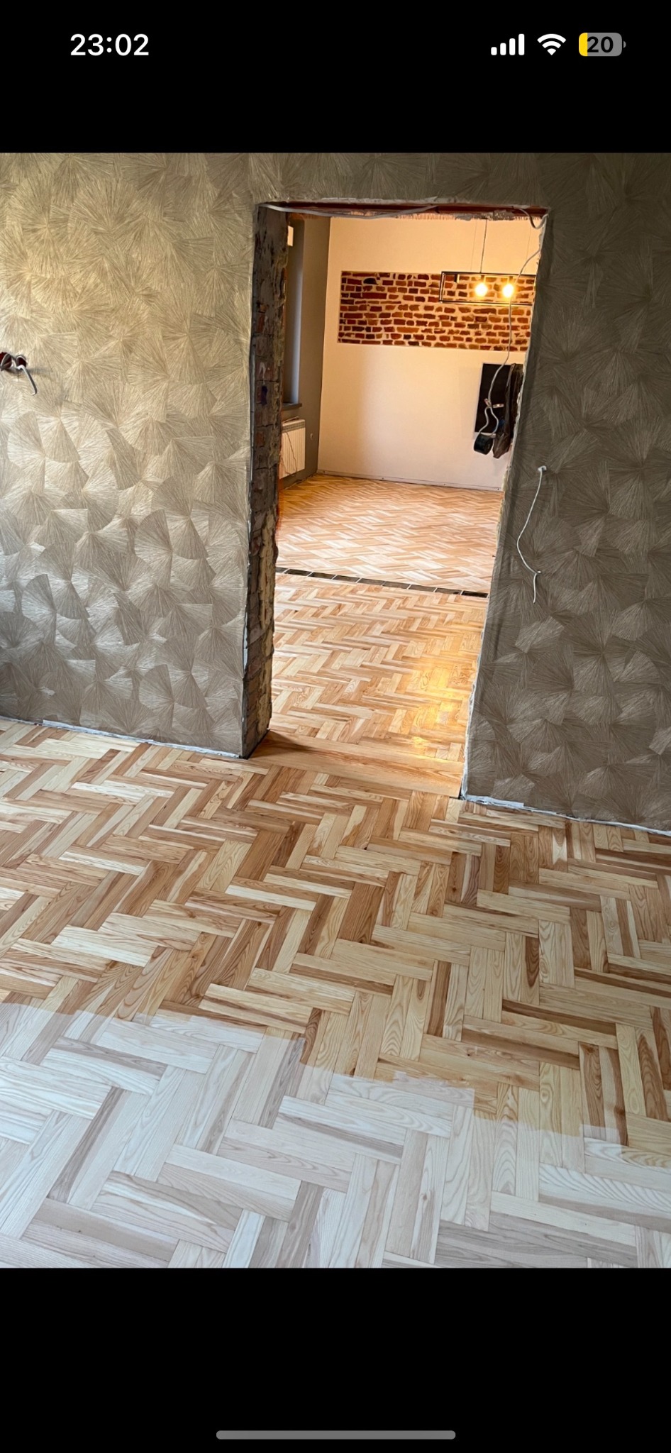 Parkiet ułożony we wzór jodełki, częściowo wycyklinowany. Widoczne przejście między starą a nową powierzchnią. W tle ściana z cegły i ozdobna tapeta.