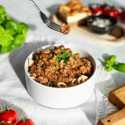 Perfect Chef Catering Dietetyczny Warszawa Dieta Pudełkowa - Porcja makaronu penne z sosem bolońskim w białej miseczce, udekorowana świeżą natką pietruszki. W tle pomidorki koktajlowe i bazylia.
