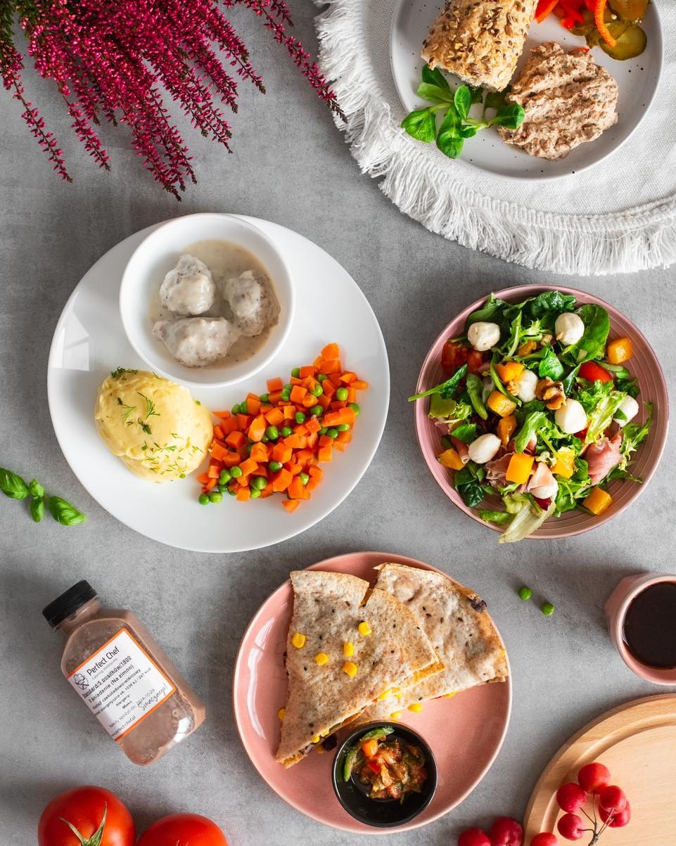 Zestaw różnorodnych dań: kotleciki w sosie, puree, warzywa, sałatka z mozzarellą, quesadilla i napój w butelce, ułożonych na talerzach na szarym tle.