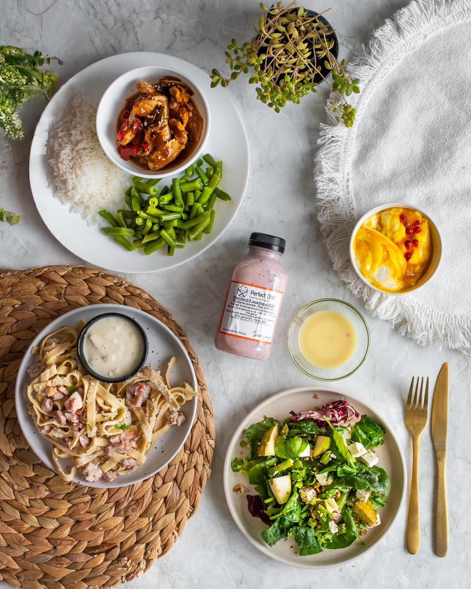 Kompozycja dań: makaron z sosem, sałatka ze szpinakiem, ryż z kurczakiem i warzywami, deser oraz butelka różowego smoothie na marmurowym tle.