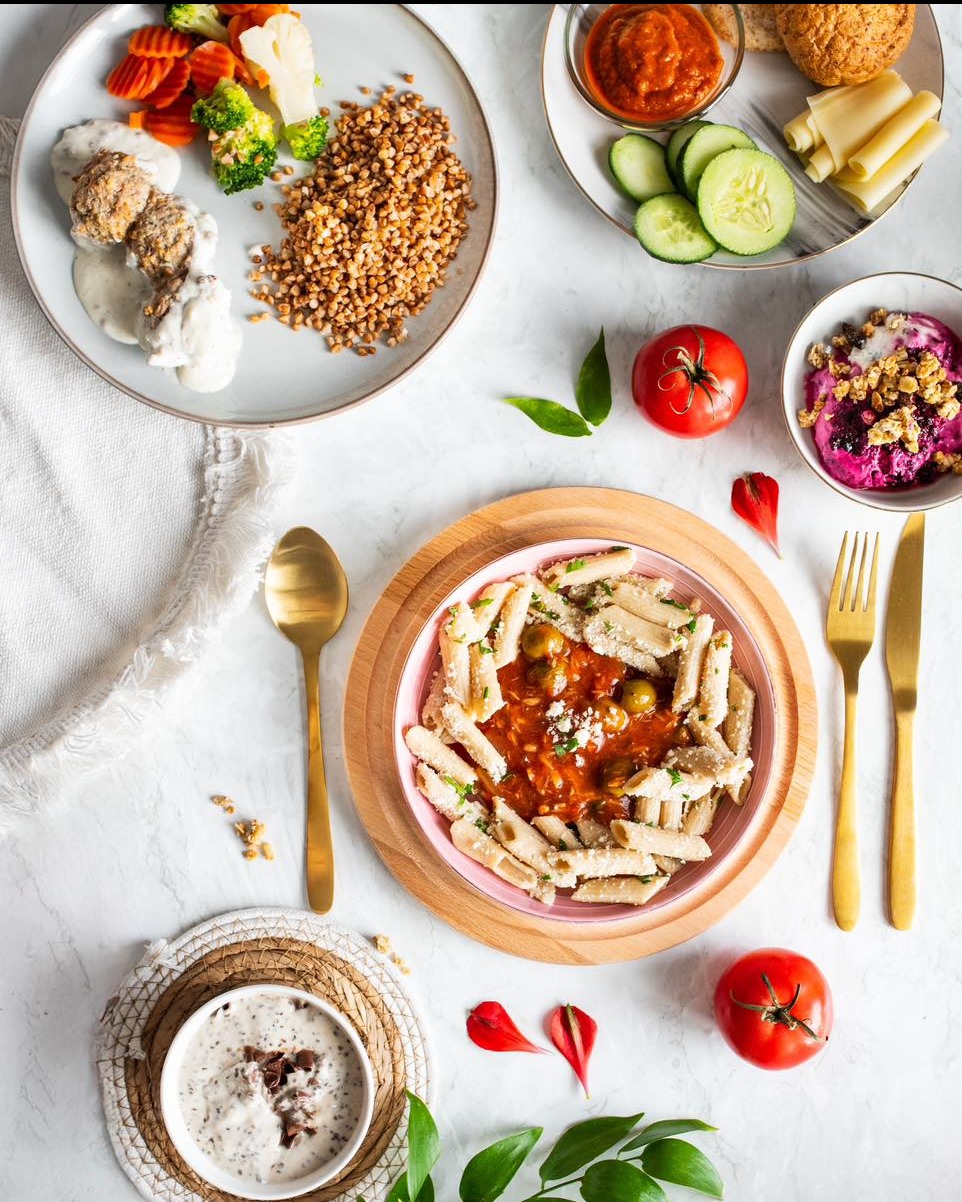 Kompozycja z różnorodnych dań: kasza gryczana, warzywa, pasta z sosem pomidorowym i deser, ułożonych na białym tle, z dodatkiem złotych sztućców i dekoracji.