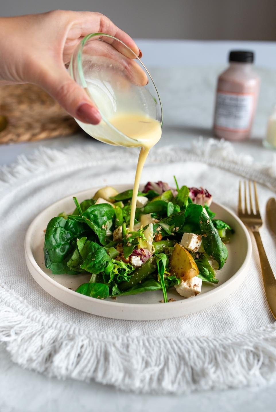 Dressing wylewany na talerz świeżej sałatki z gruszką, serem i orzechami; w tle butelka z sosem i złote sztućce na białym obrusie z frędzlami.