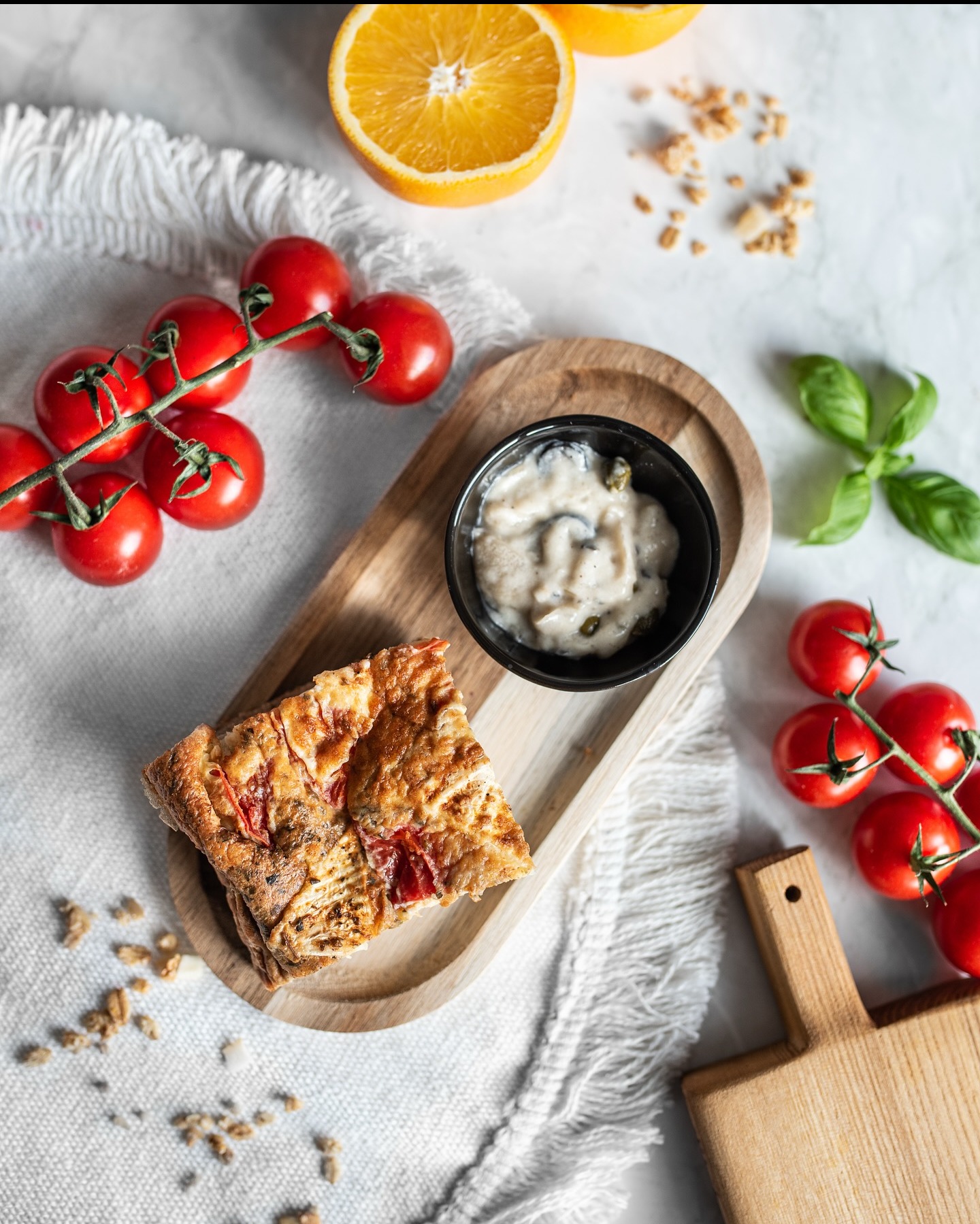 Kawałek tarty na drewnianej desce z sosem, pomidorkami cherry, pomarańczą i bazylią na białym obrusie. Zdjęcie z góry, naturalne światło.