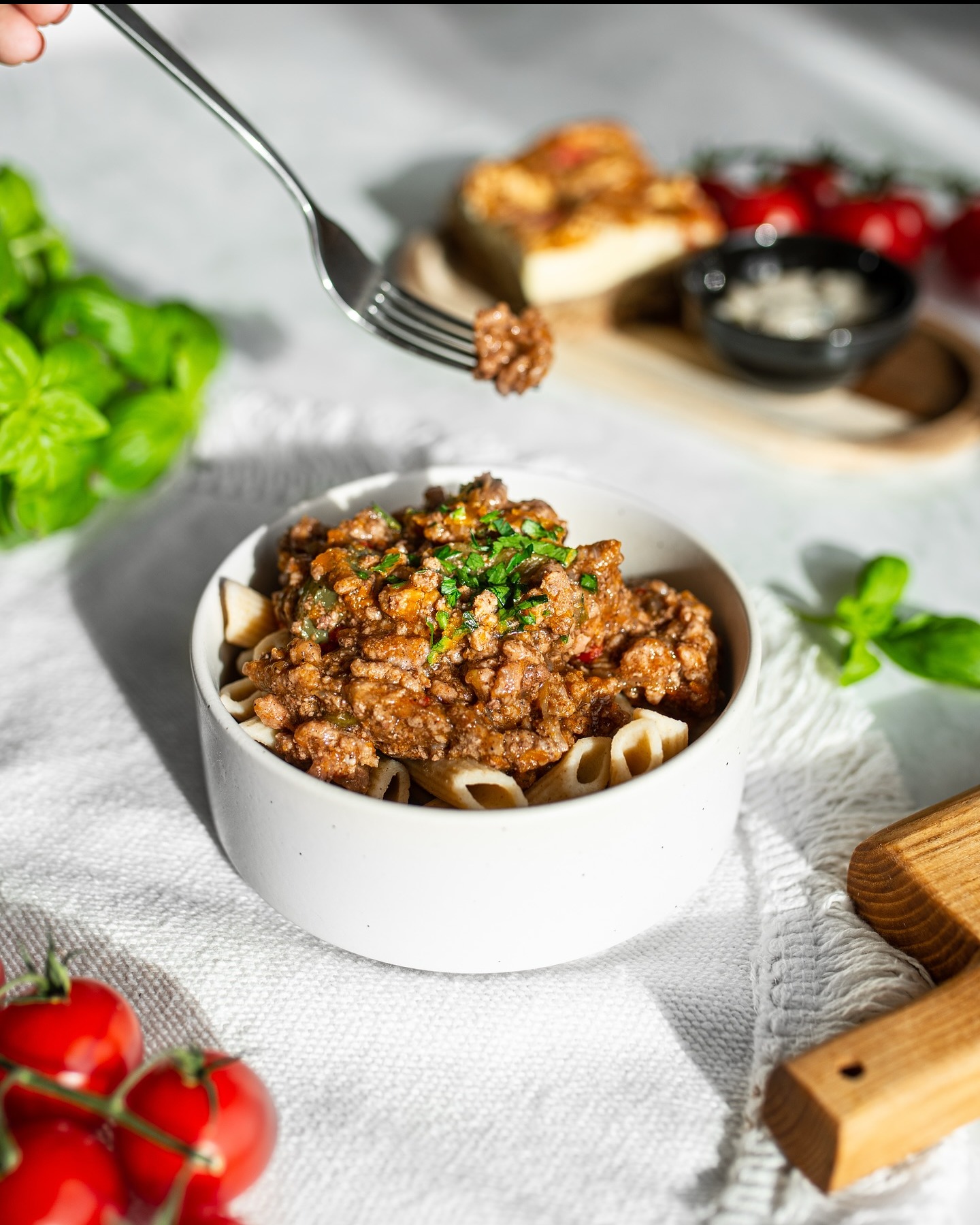 Porcja makaronu penne z sosem bolońskim w białej miseczce, udekorowana świeżą natką pietruszki. W tle pomidorki koktajlowe i bazylia.