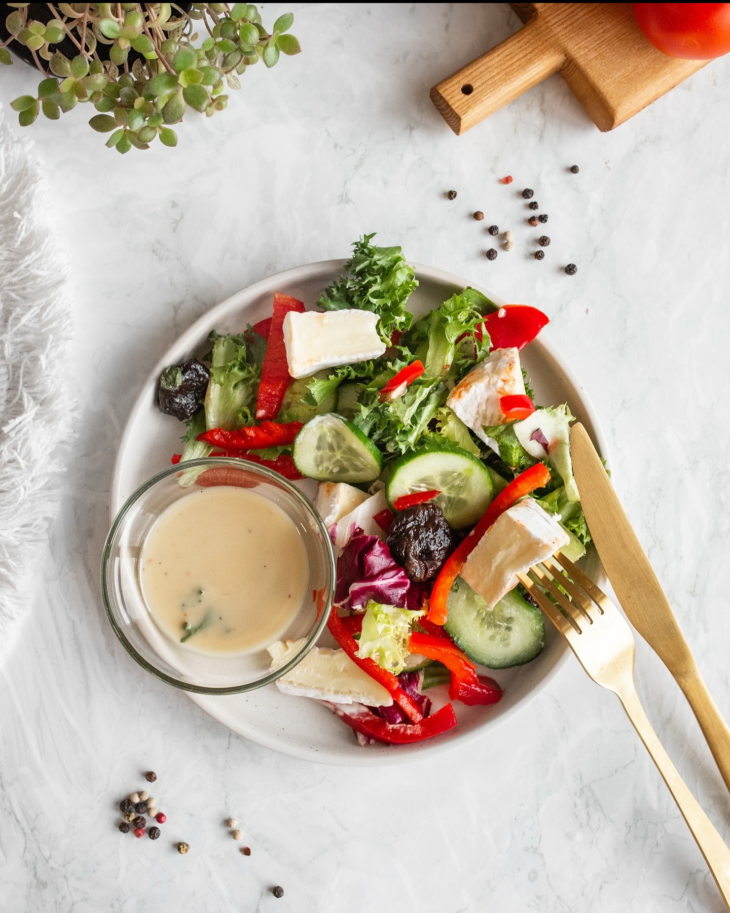 Świeża sałatka z serem camembert, czerwoną papryką i ogórkiem, z sosem w szklanej miseczce na marmurowym tle, z dodatkiem złotych sztućców i przypraw.