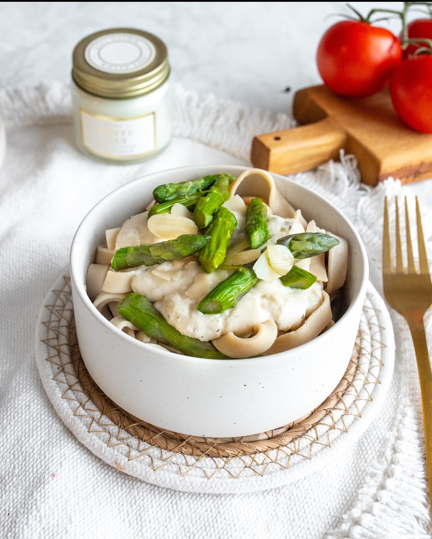 Makaron tagliatelle z sosem śmietanowym i szparagami w białej misce na plecionym podkładce. W tle pomidory i świeca.