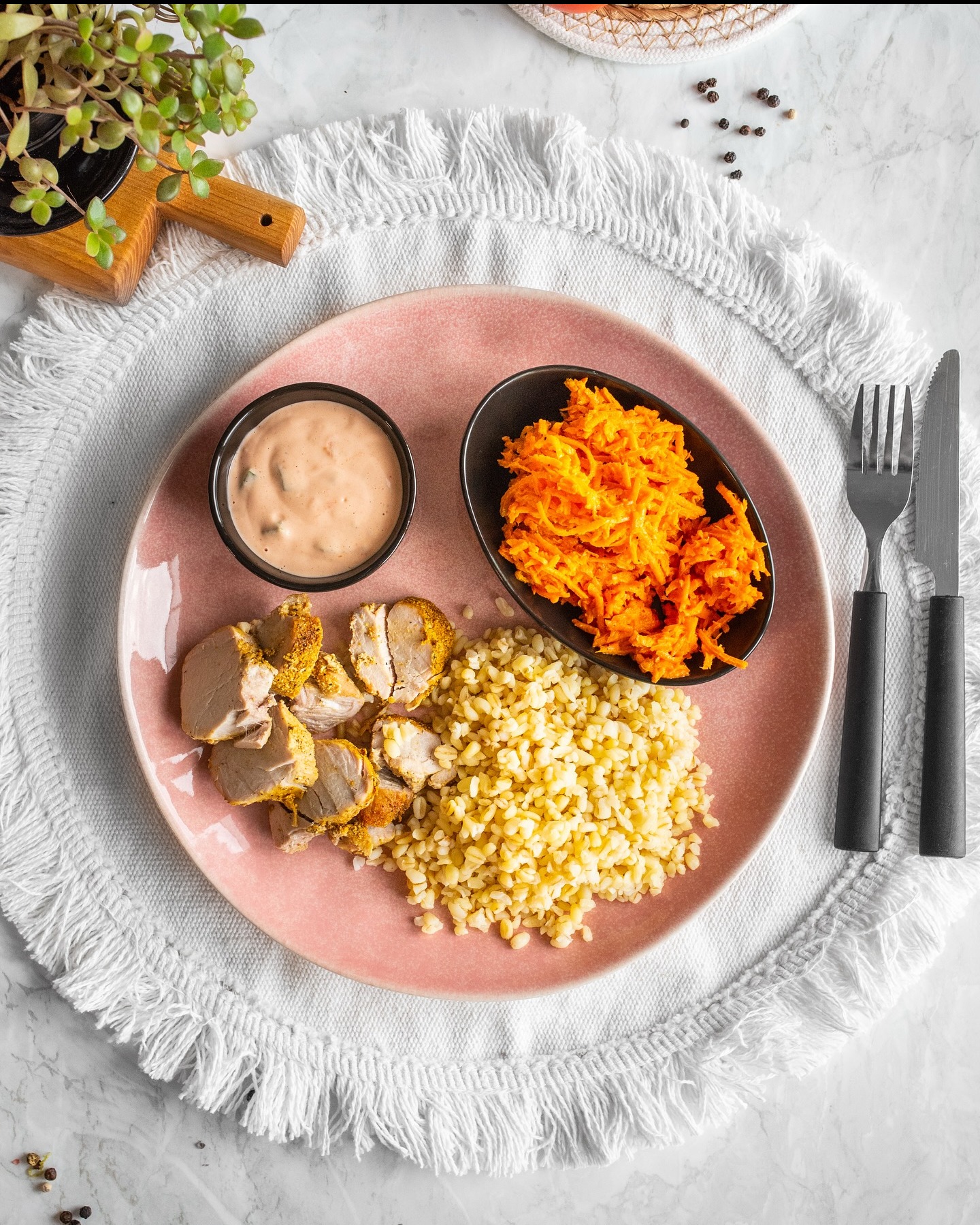 Różowy talerz z mięsem, kaszą bulgur, marchewką i sosem na białym obrusie z frędzlami, ułożony na marmurowym blacie z przyprawami i zieloną rośliną.