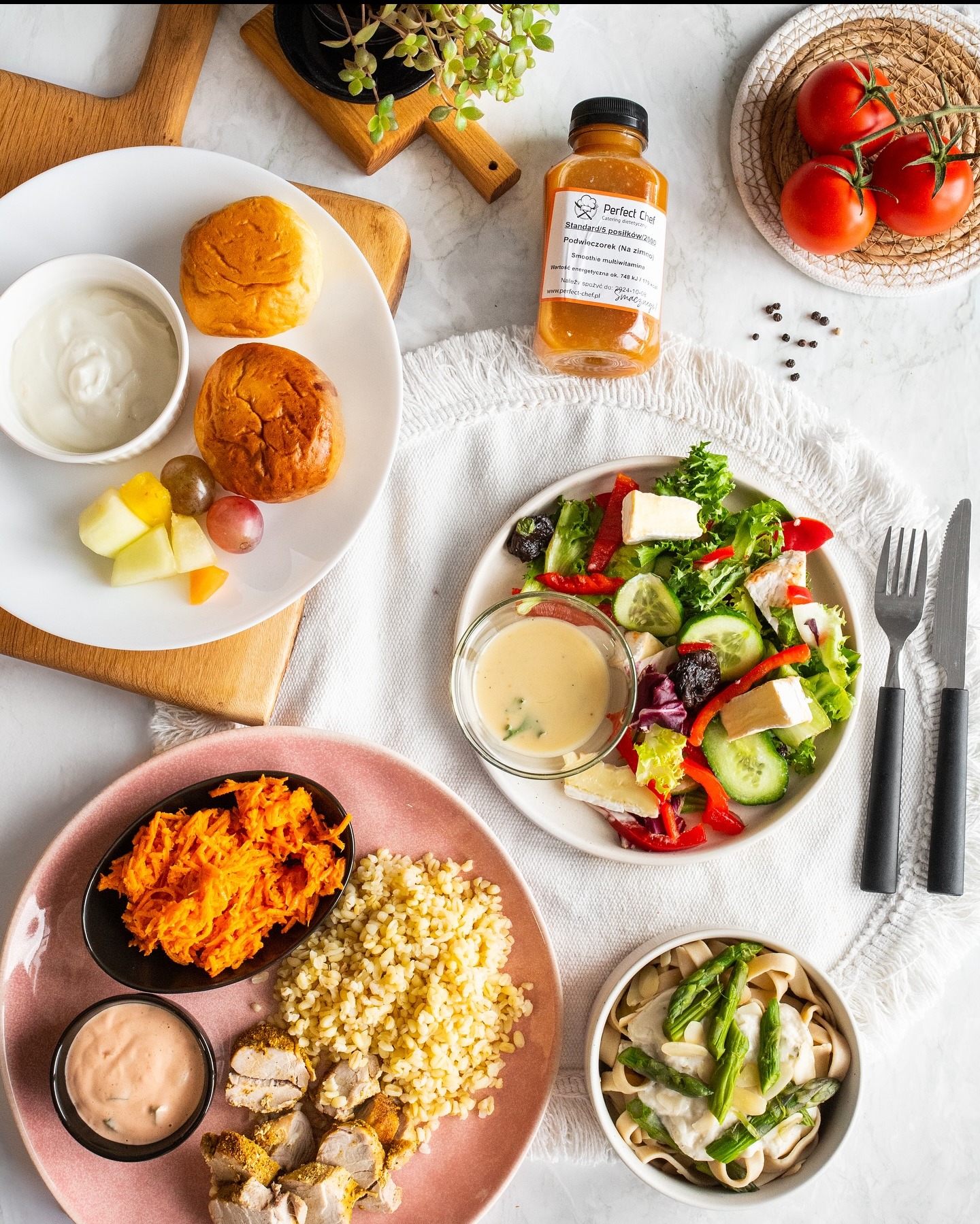 Zestaw posiłków dietetycznych: sałatka z serem, pasta z warzywami, smoothie, pieczywo i owoce na białym stole z dekoracją.