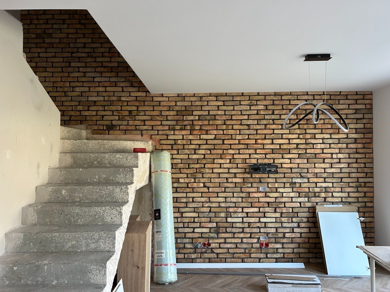 Ceglana ściana w salonie z surowymi schodami, nowoczesna lampa wisząca. Widoczne puszki elektryczne i elementy wykończeniowe. Parkiet ułożony we wzór jodełki.