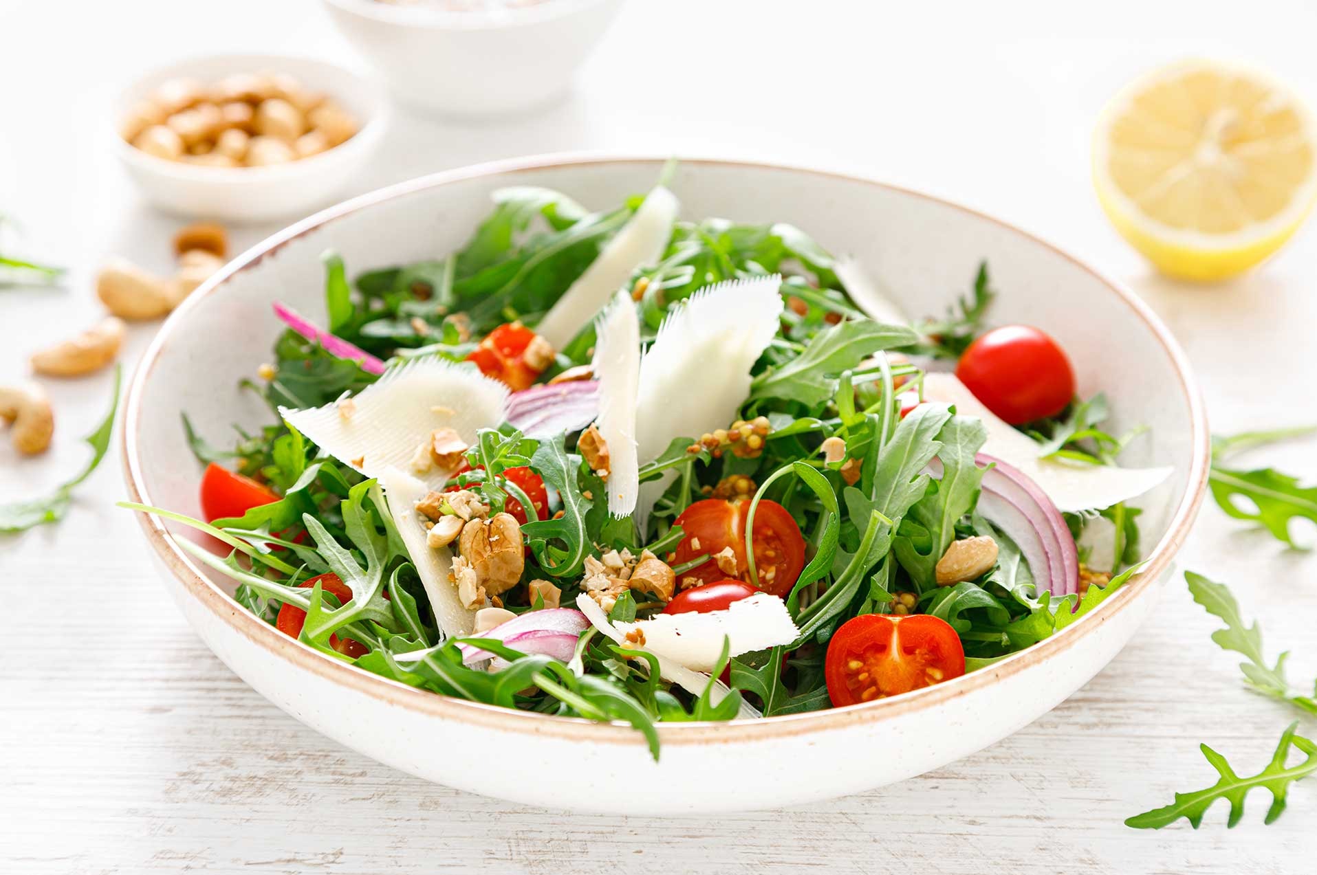 Świeża sałatka z rukoli, pomidorków cherry, czerwonej cebuli, parmezanu i orzechów w białej misce, z cytryną i orzechami w tle, na białym drewnianym stole.