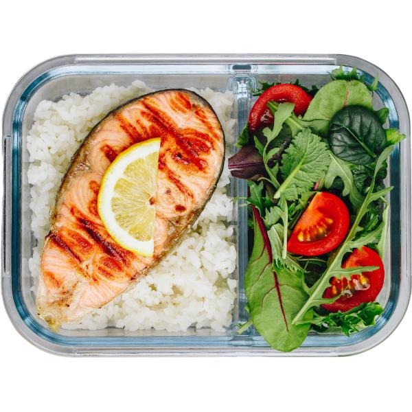 Lunchbox z grillowanym łososiem, ryżem i sałatką z pomidorami i zieleniną. Plasterek cytryny na rybie. Transparentne pudełko z przegródkami.