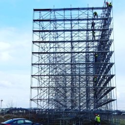 DELTA-BUD - Montaż rusztowania modułowego na tle błękitnego nieba. Na konstrukcji widoczni pracownicy w odblaskowych ubraniach. U dołu czarny samochód osobowy.